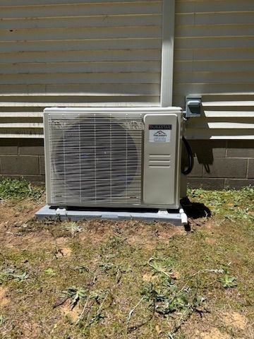 Outdoor air conditioner unit beside a house wall, with a hose attached on a grassy yard.