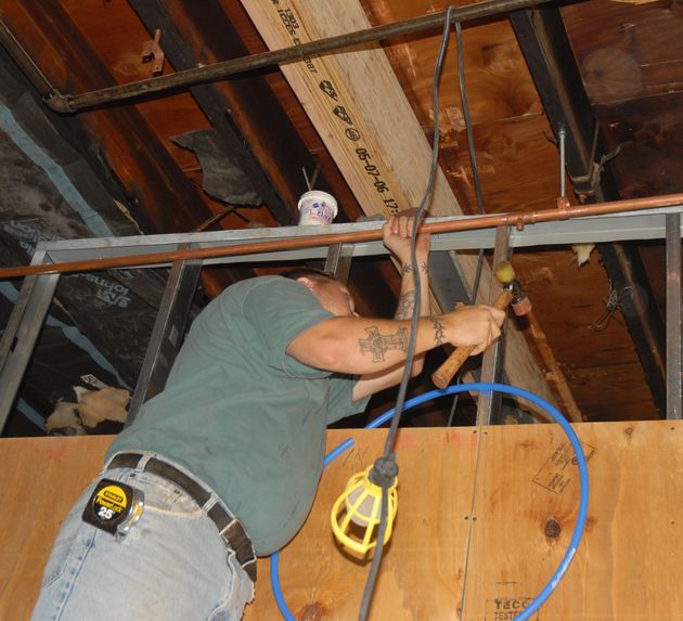 Worker in a hard hat wiring ceiling beams in an unfinished room, holding tools and blue cable.