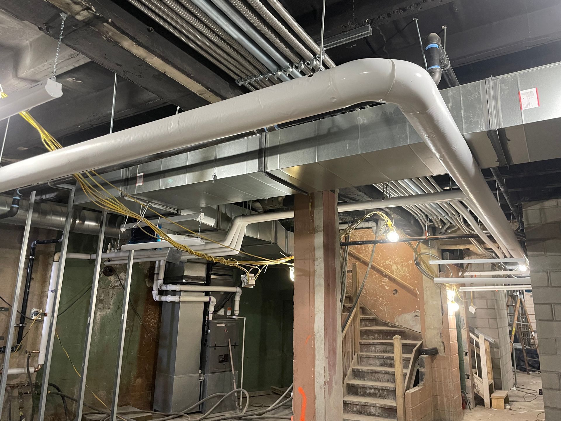 Industrial basement with exposed pipes, ducts, and stairway beside brick walls and concrete columns