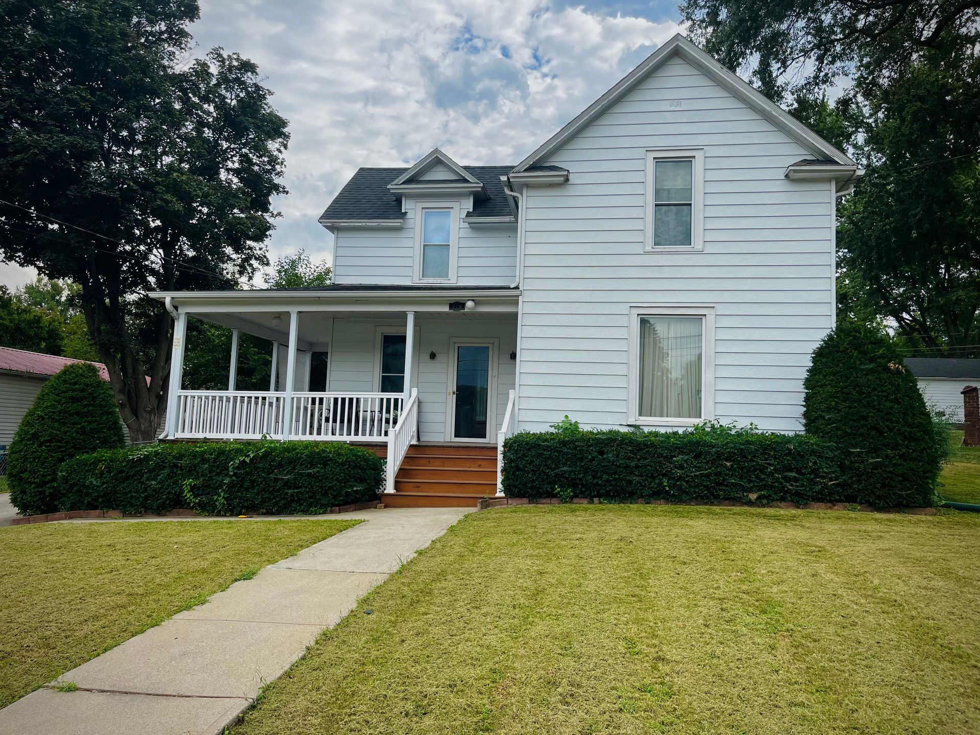 White two-story house with front porch, shrubs, and a sidewalk leading across a green lawn