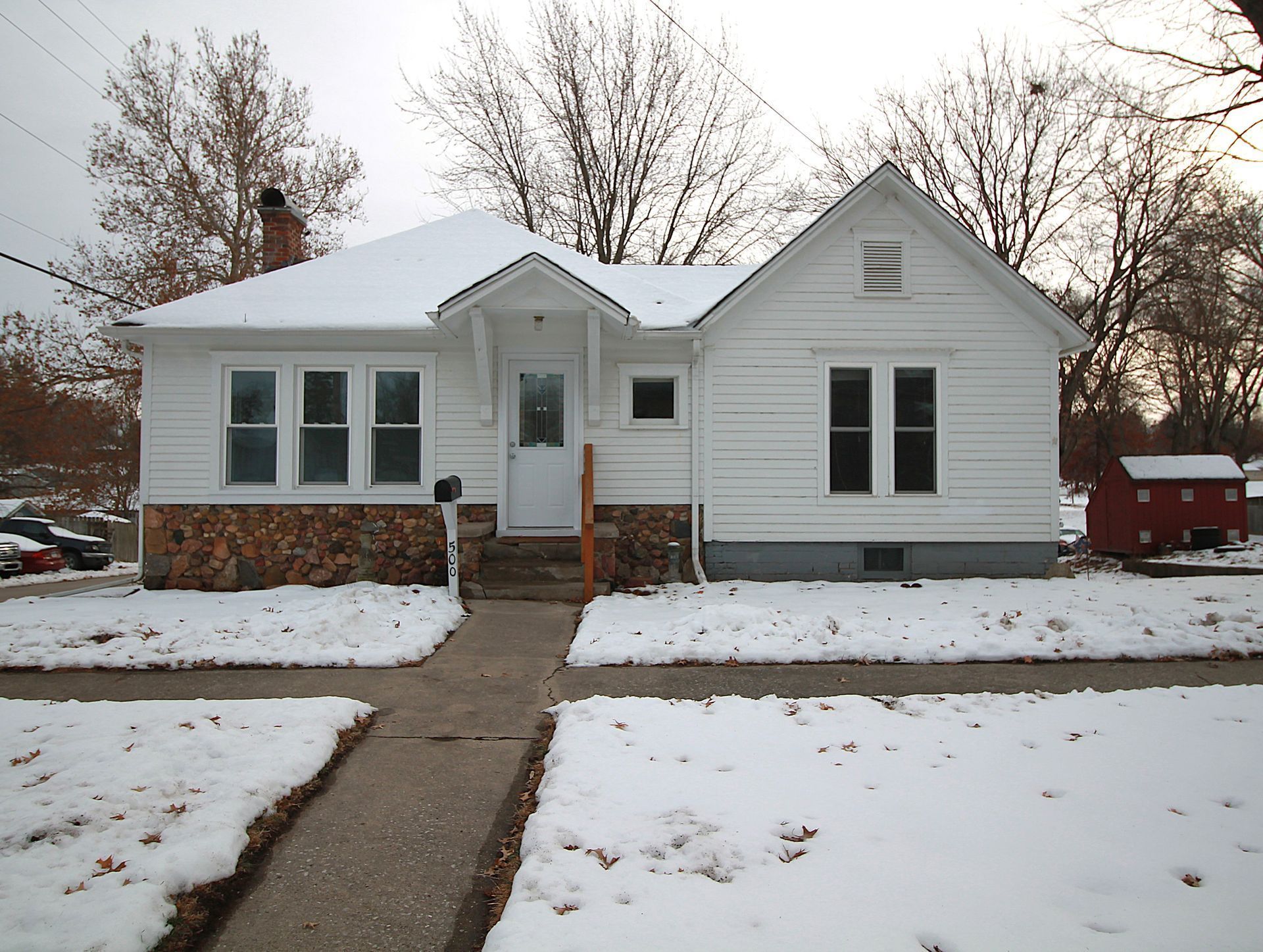 Small white house with snow-covered roof, stone foundation, and sidewalk.