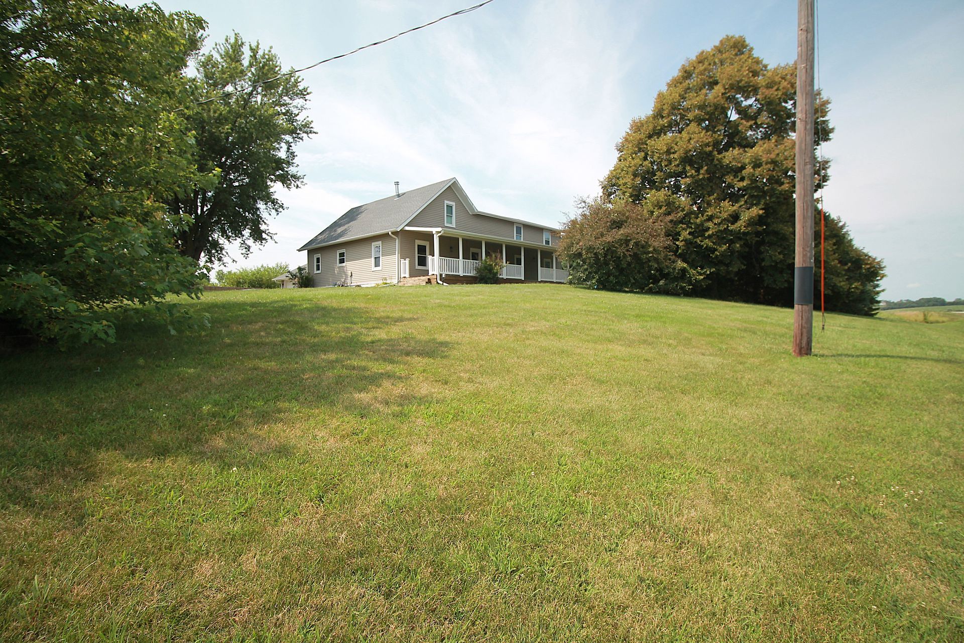 Small white house on a grassy hill with trees and a utility pole under a cloudy sky