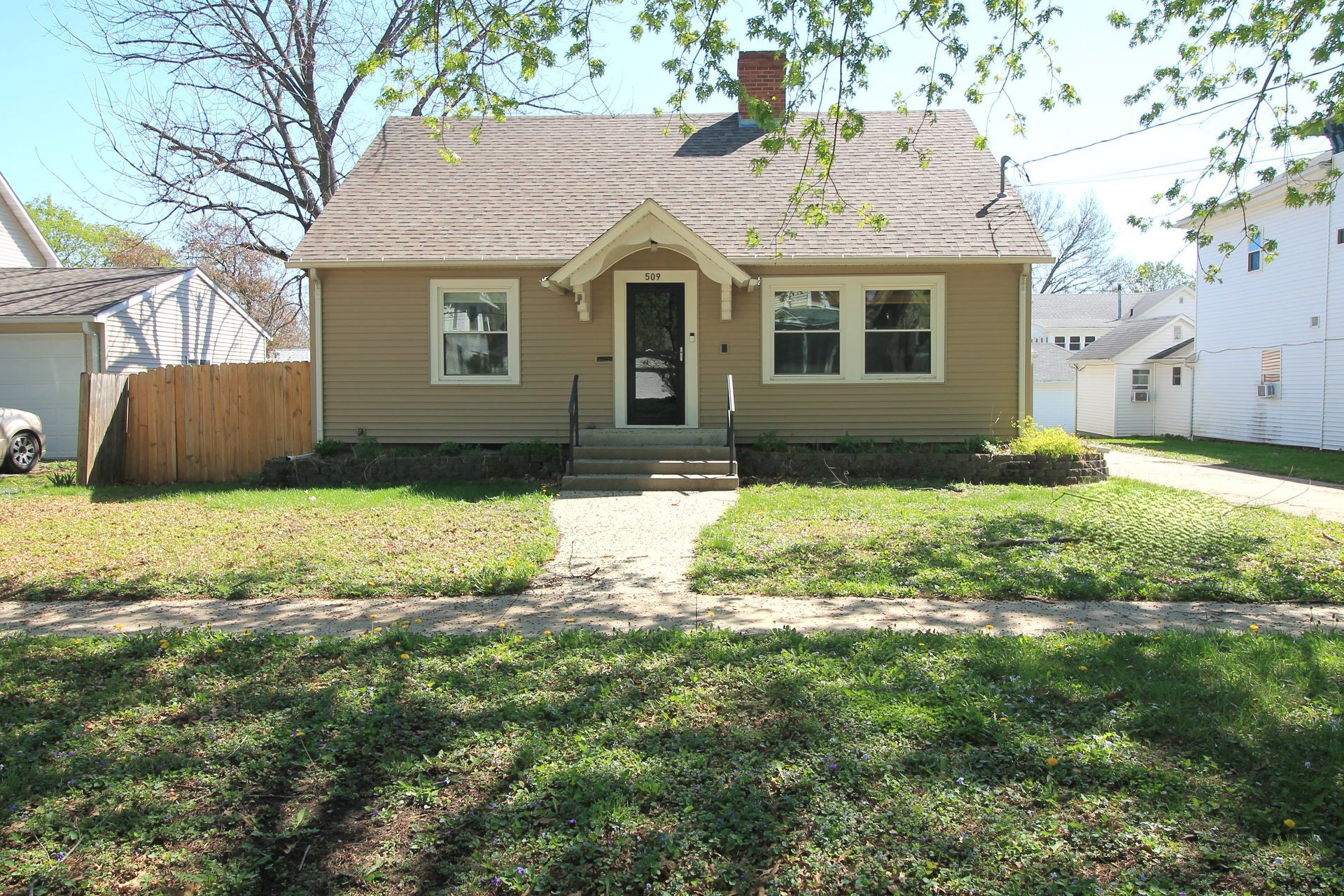 Small beige house with front porch, white-trimmed windows, and a grassy yard under trees