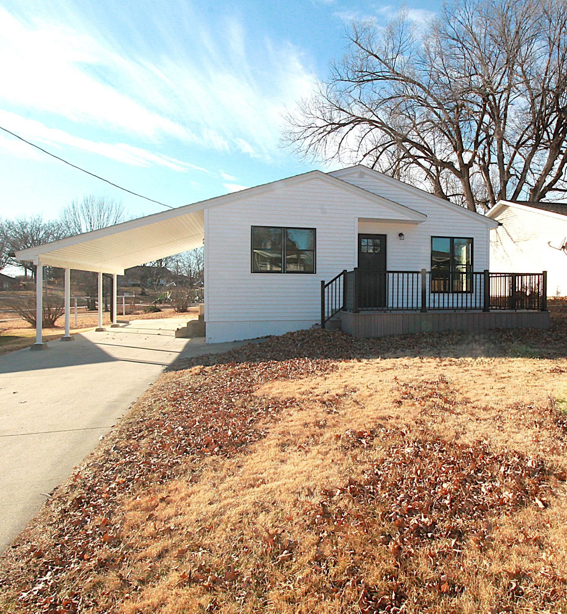 White house with carport, black door and windows, sits on a slight hill with brown grass.
