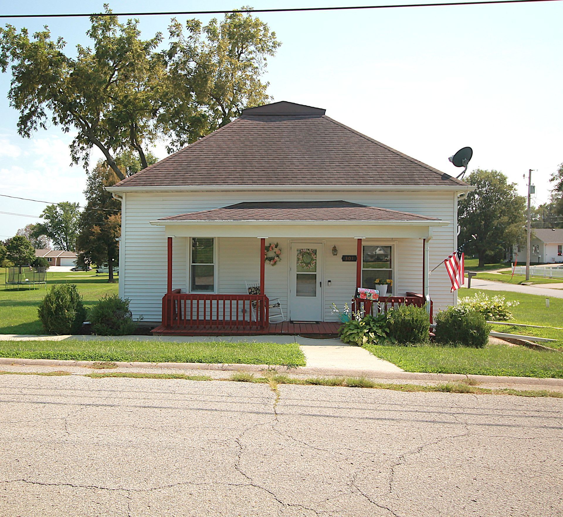 Small white house with a dark roof, front porch, and American flag beside a quiet street