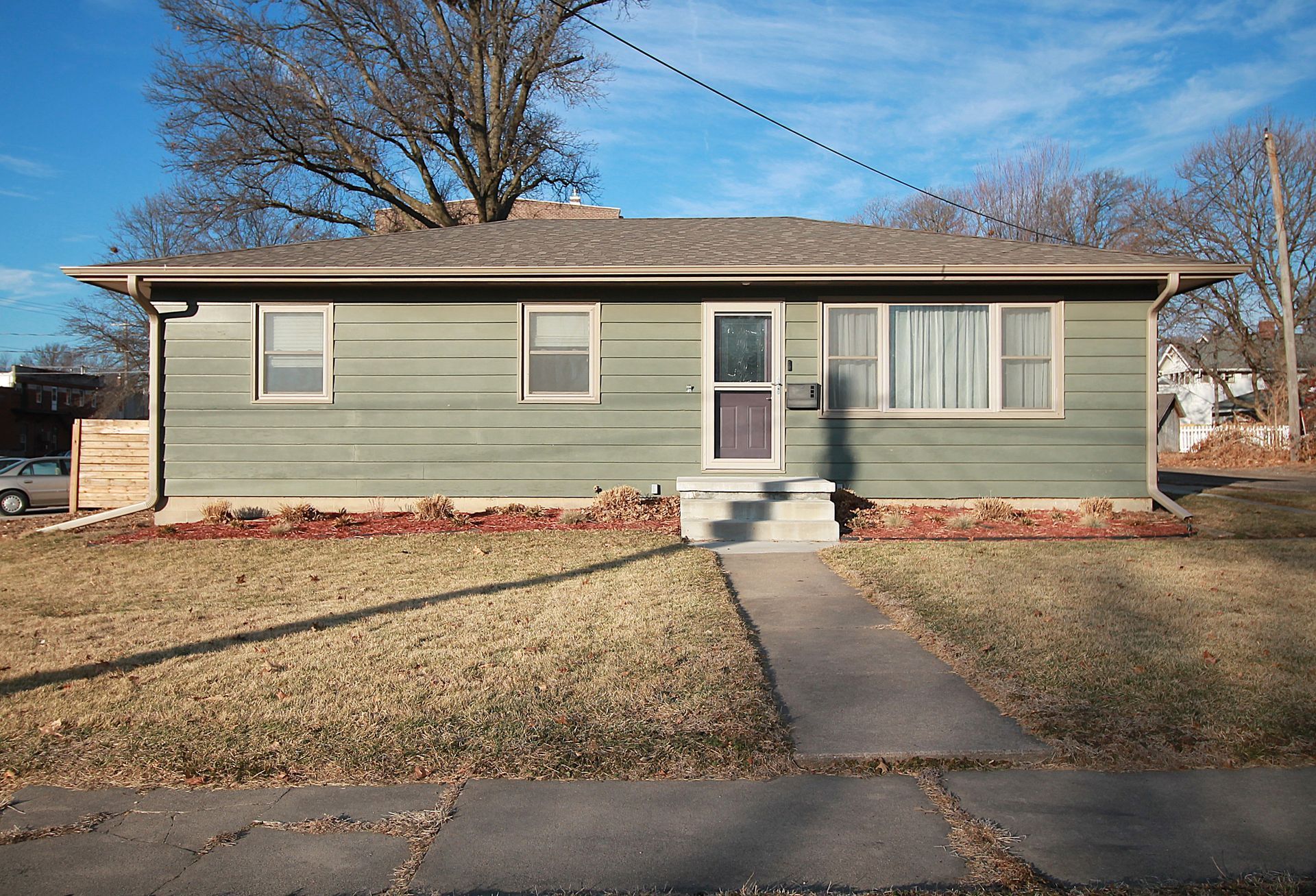 White house with carport, black door and windows, sits on a slight hill with brown grass.