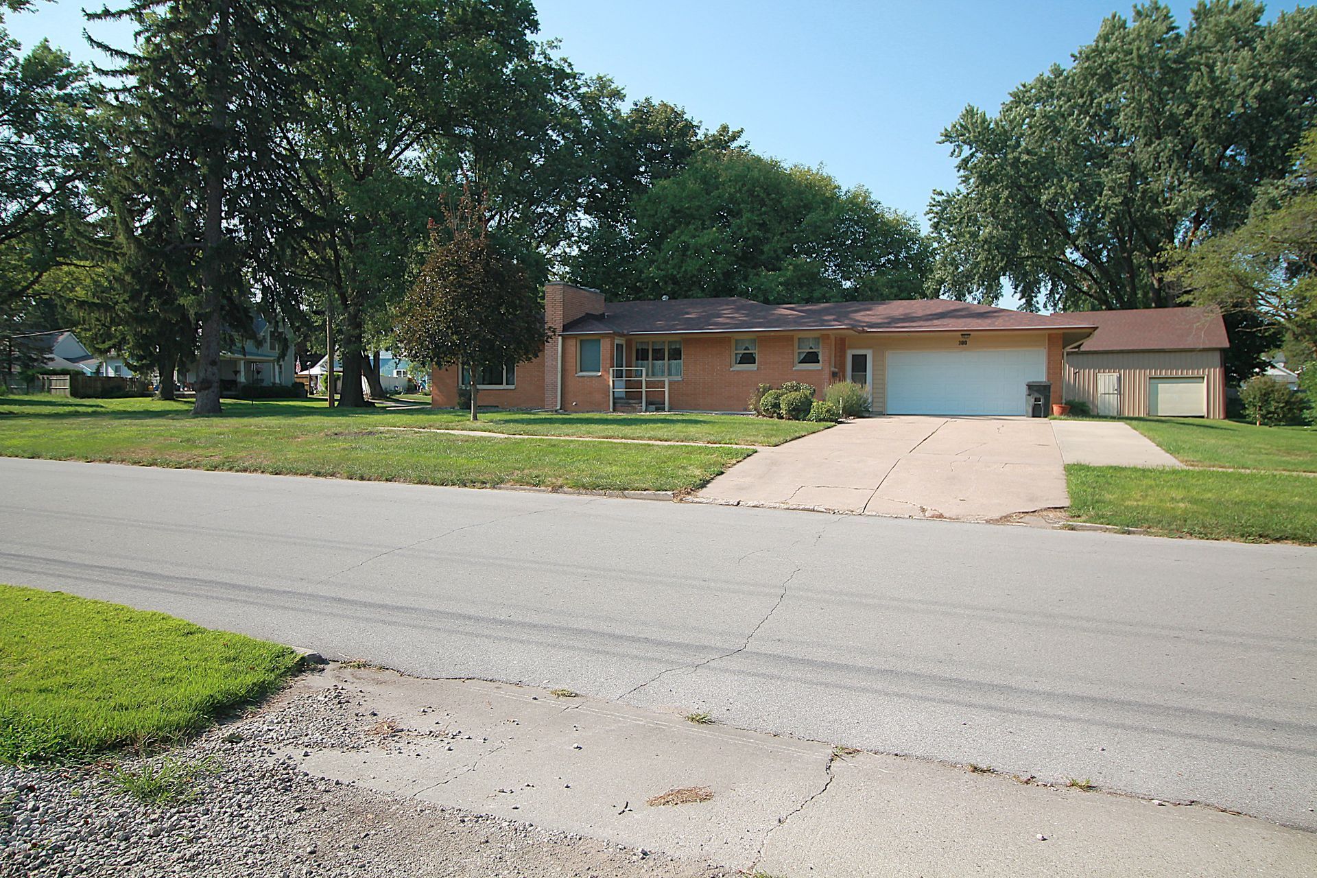 Suburban brick house with a wide driveway, garage, and large trees along a quiet street