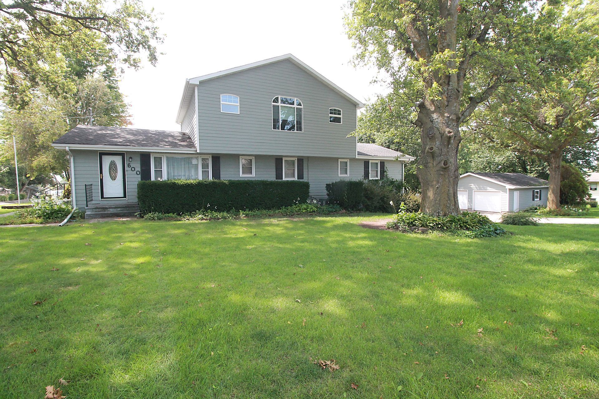 Suburban two-story house with gray siding, green lawn, and large trees in the yard.