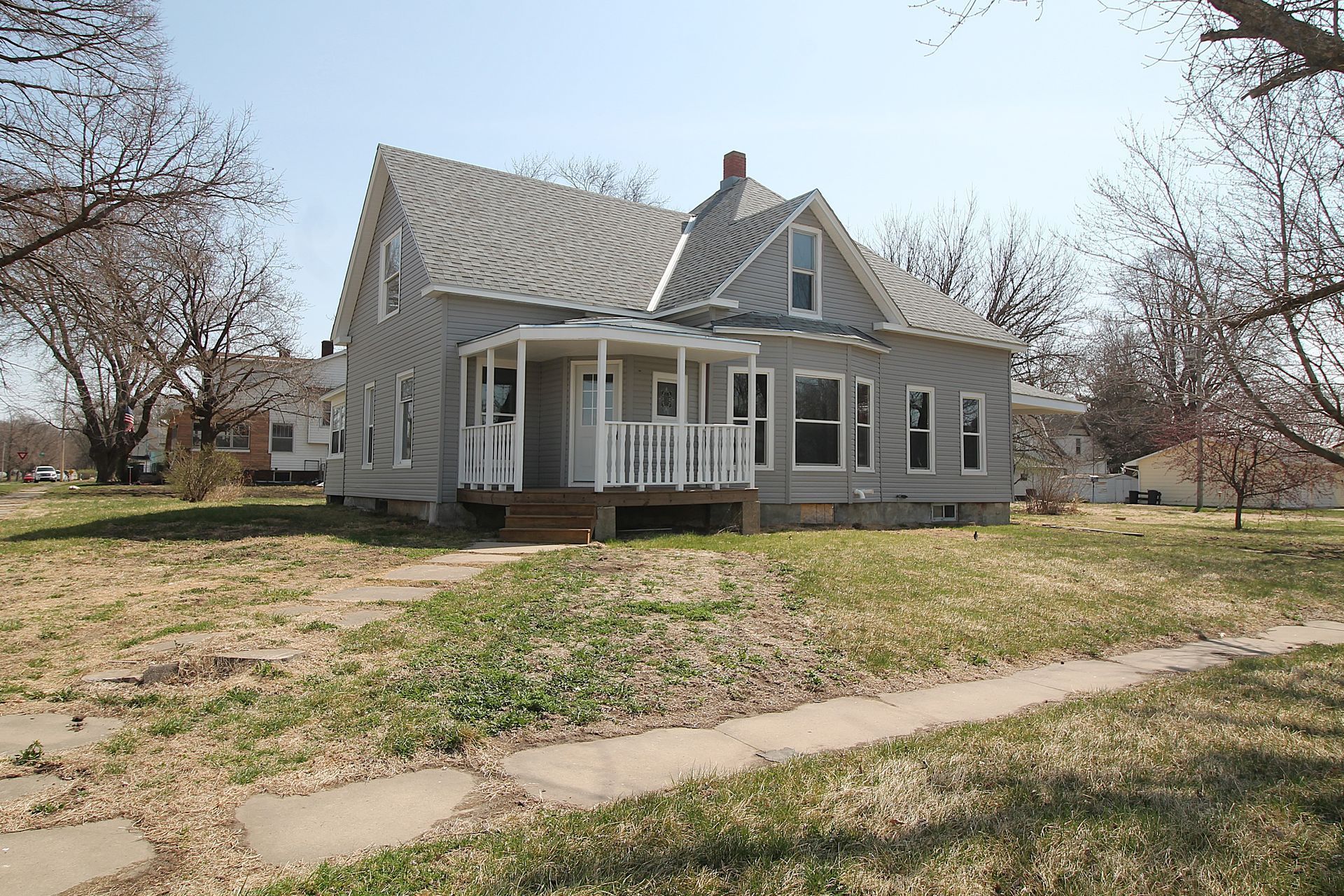 Gray farmhouse with a wraparound porch on a grassy lot, viewed from the front under bare trees