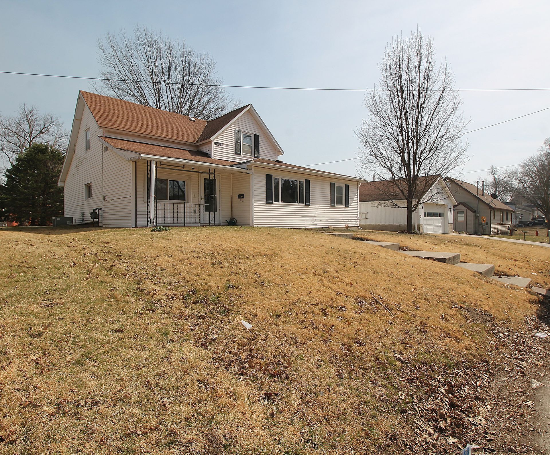 White house with red roof on a grassy hill, leafless trees, and a driveway on a cloudy day