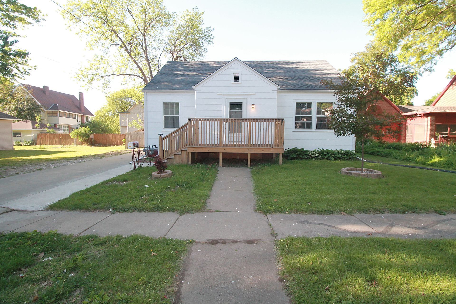 White house with a wooden front porch and steps, centered on a sidewalk in a leafy suburban yard.