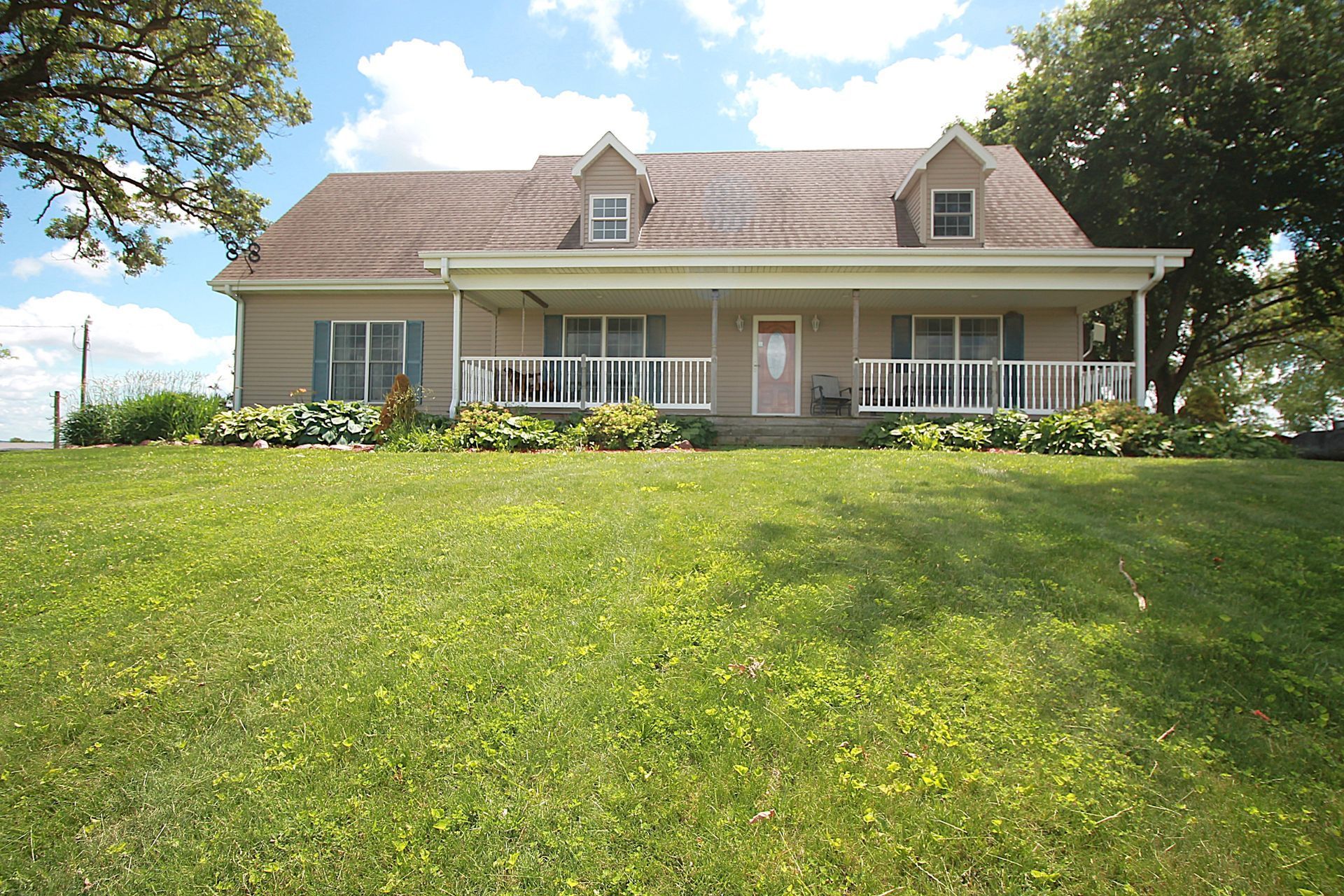 Large beige house with a porch and dormer windows on a grassy hill under a partly cloudy sky