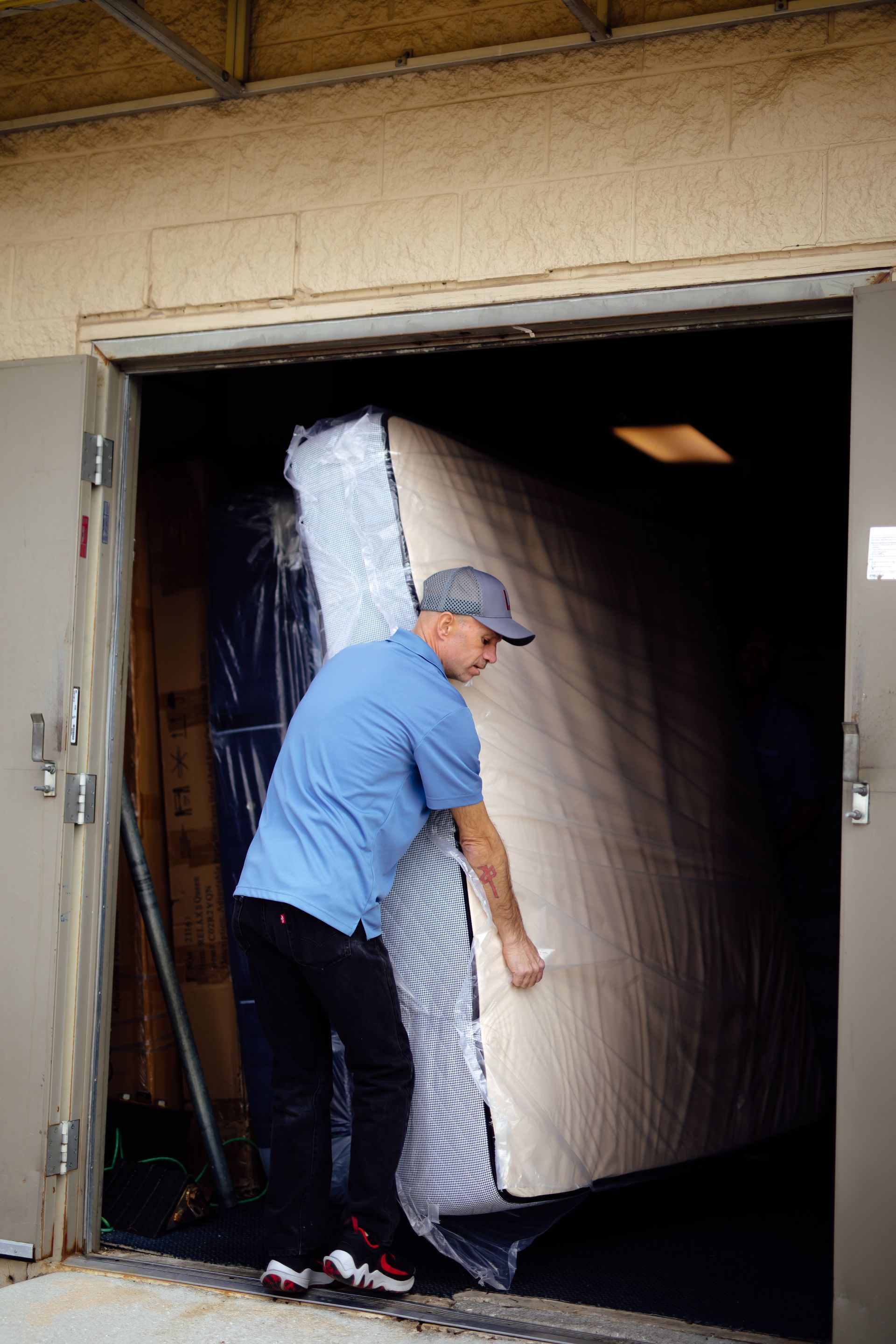 Person in a blue shirt and cap moving a large mattress through a doorway into a garage