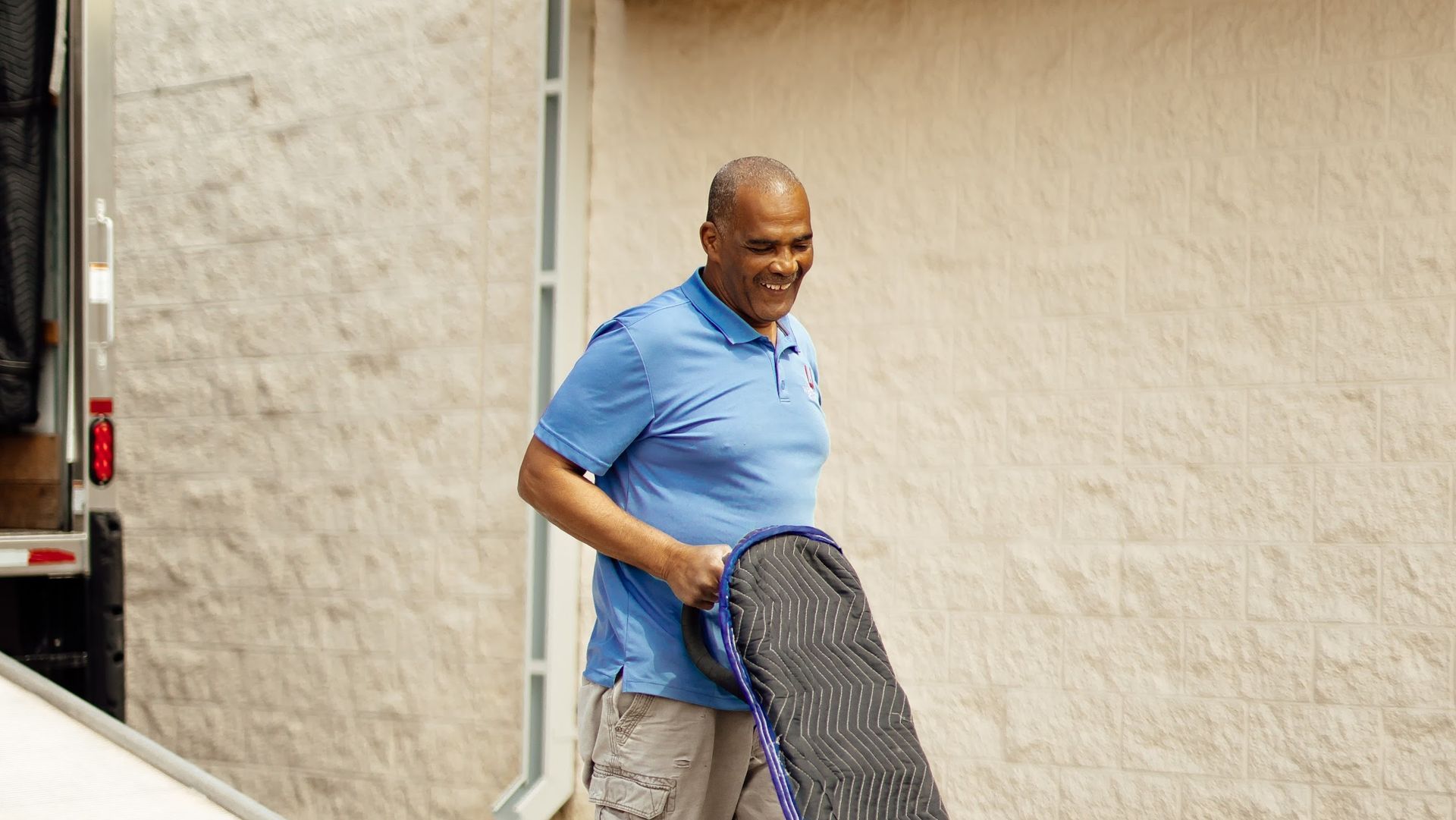 Man in blue polo carrying a large black bag beside a truck near a beige wall