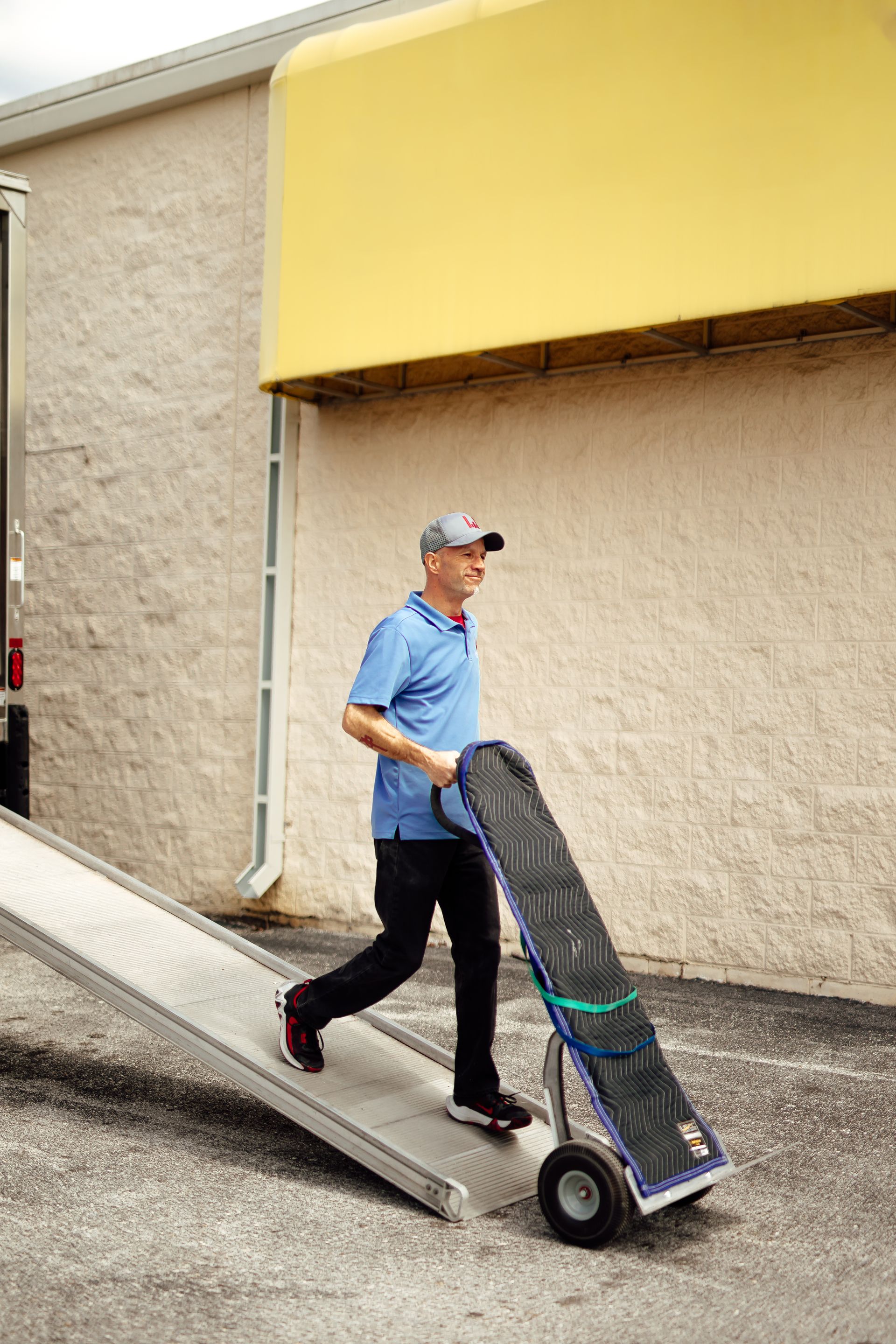 Man pushing a dolly down a ramp beside a beige building with a yellow awning