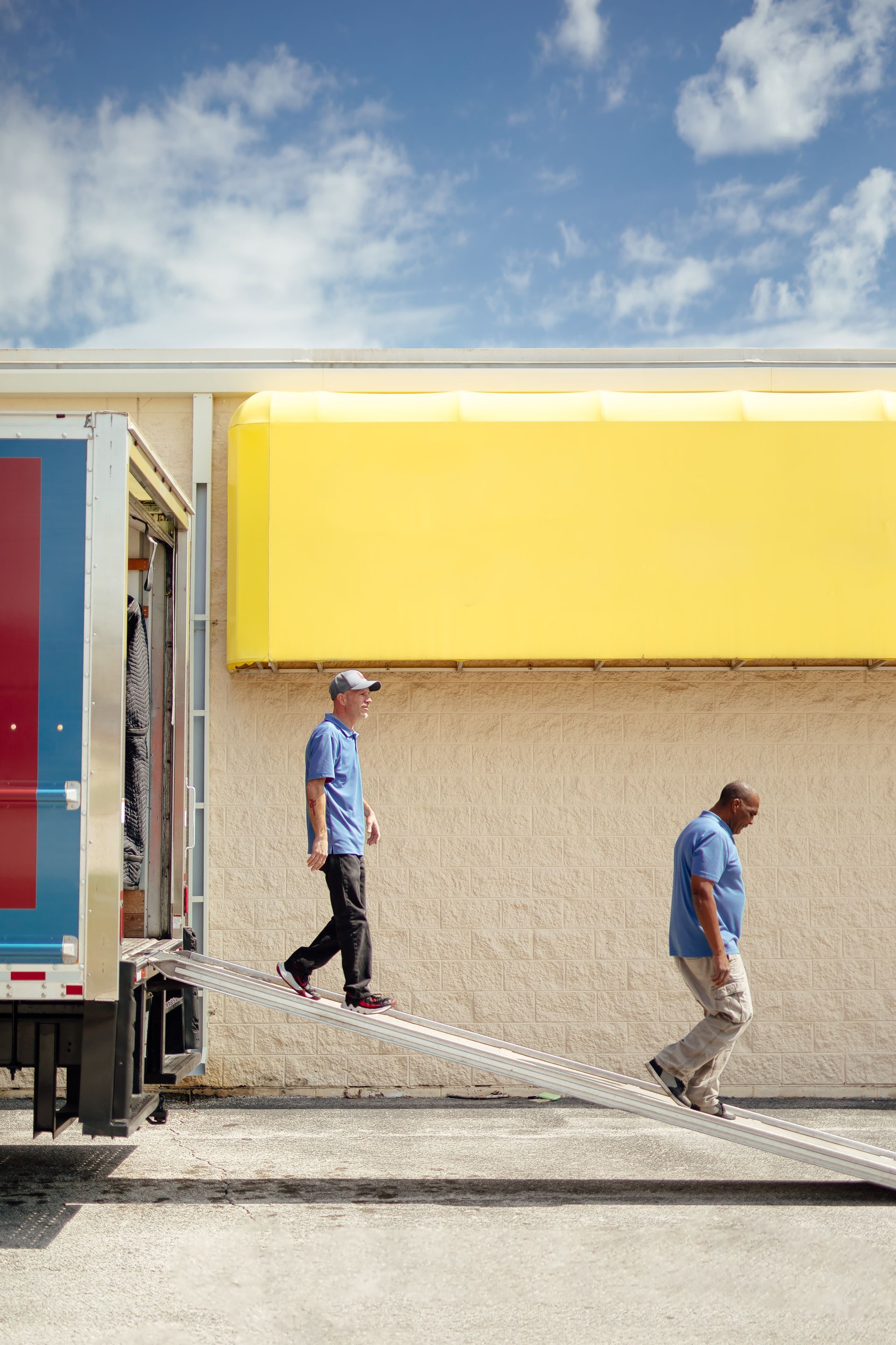 Two workers unload a truck beside a yellow building under a blue sky.
