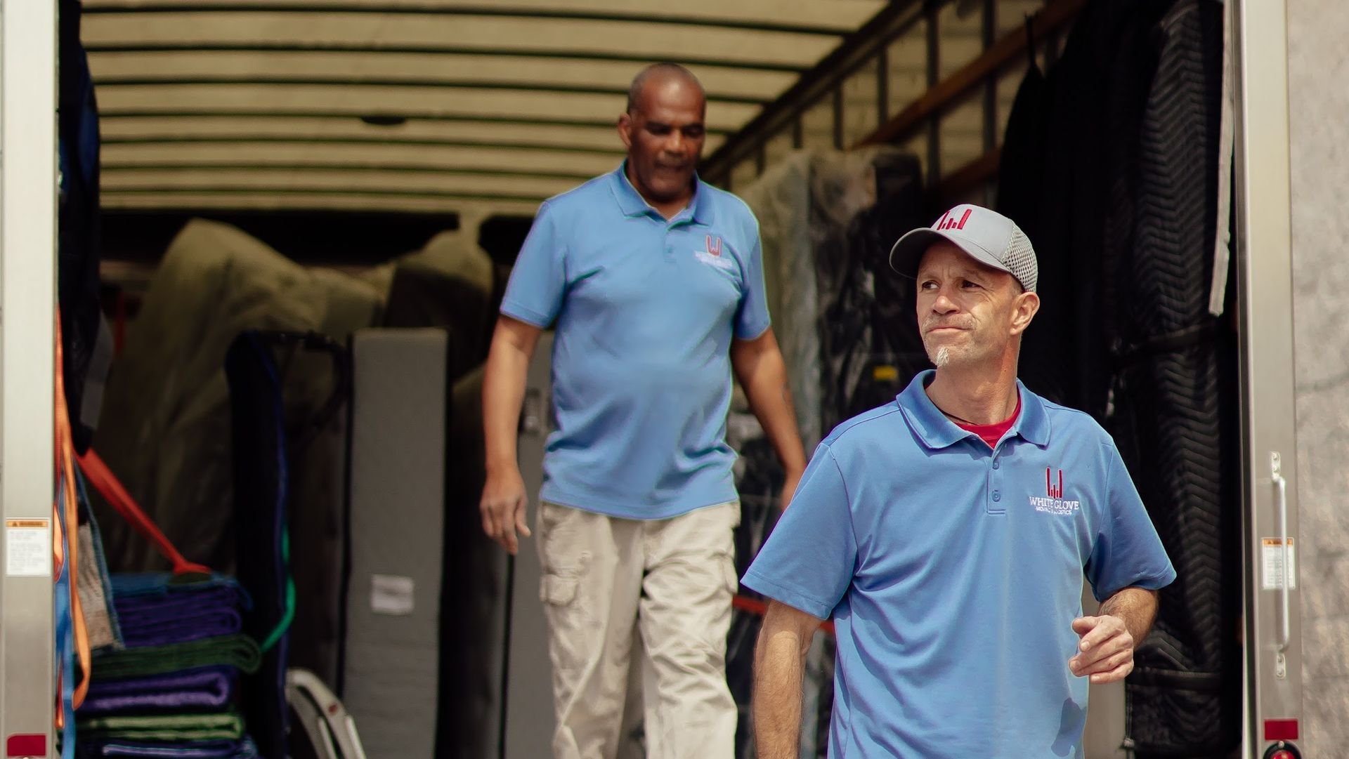 Two workers in blue polos standing in a warehouse doorway, one in a cap smiling in front.