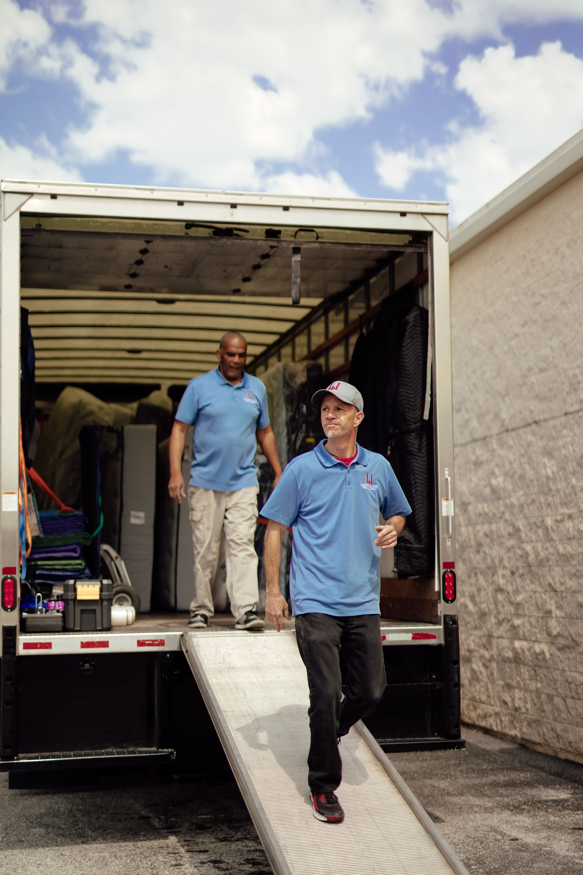 Two movers in blue shirts unloading equipment from a truck with a ramp at a building.