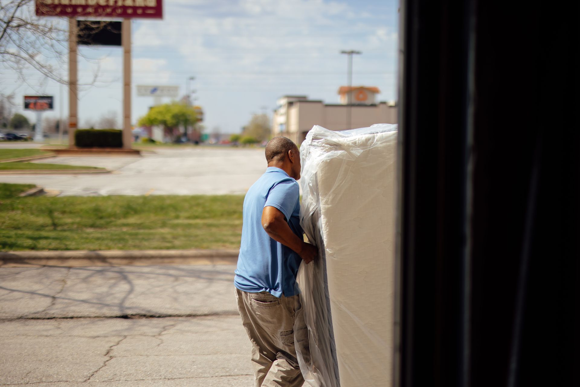 Man in blue shirt pushing a large white-wrapped item outdoors near a street.