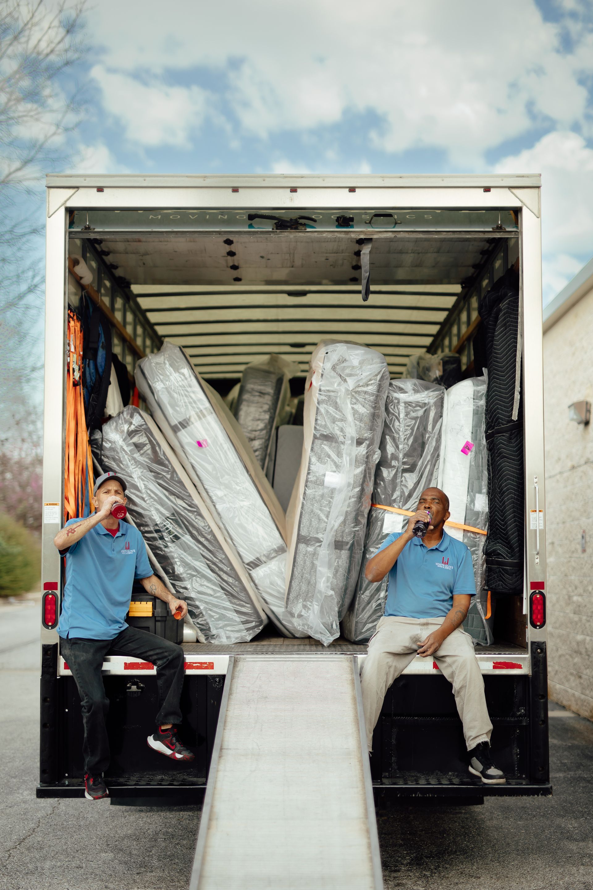 Two movers unload wrapped furniture from a truck using a metal ramp.