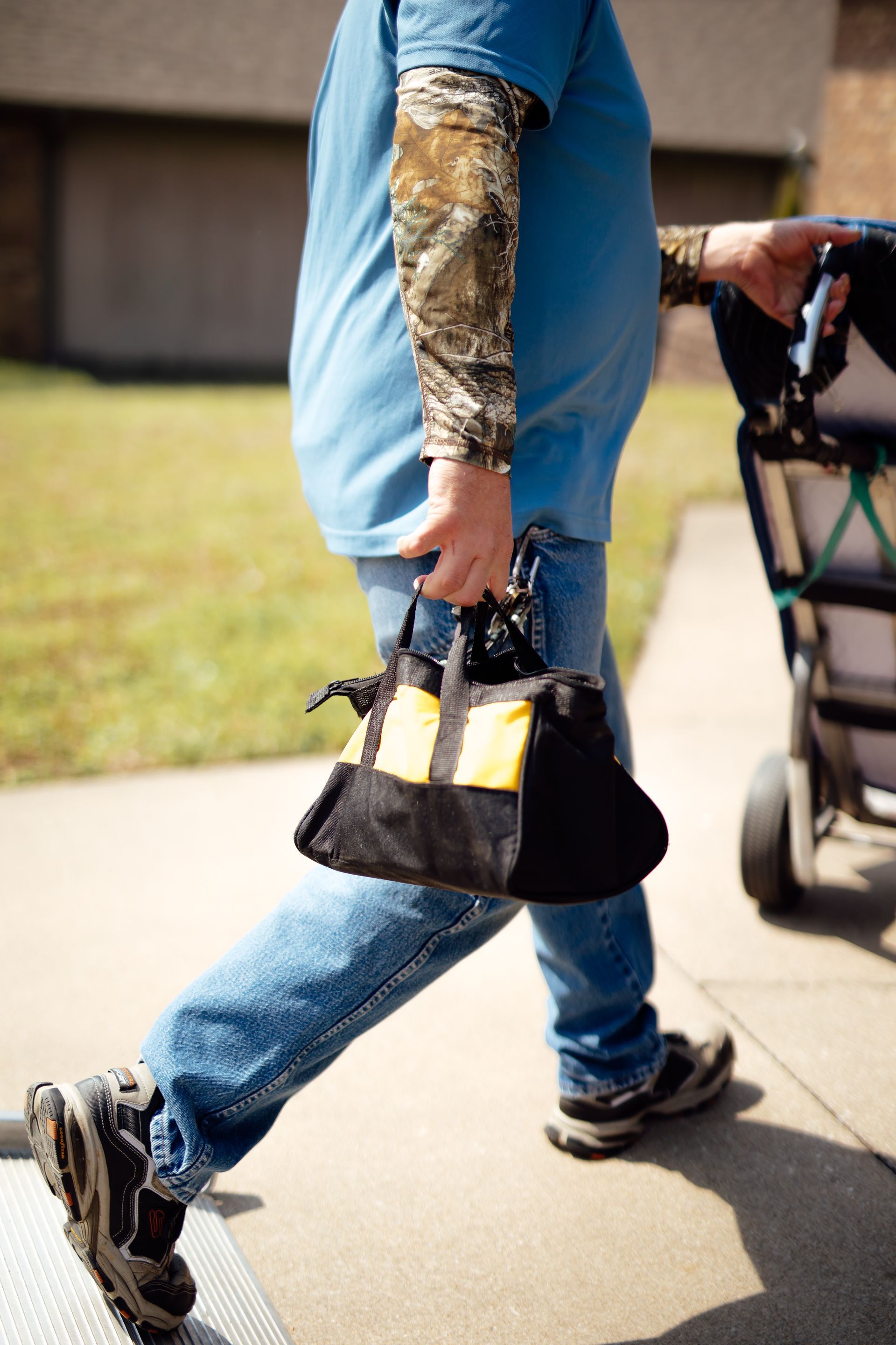 Person with a camouflaged prosthetic arm walks beside a stroller on a sidewalk.