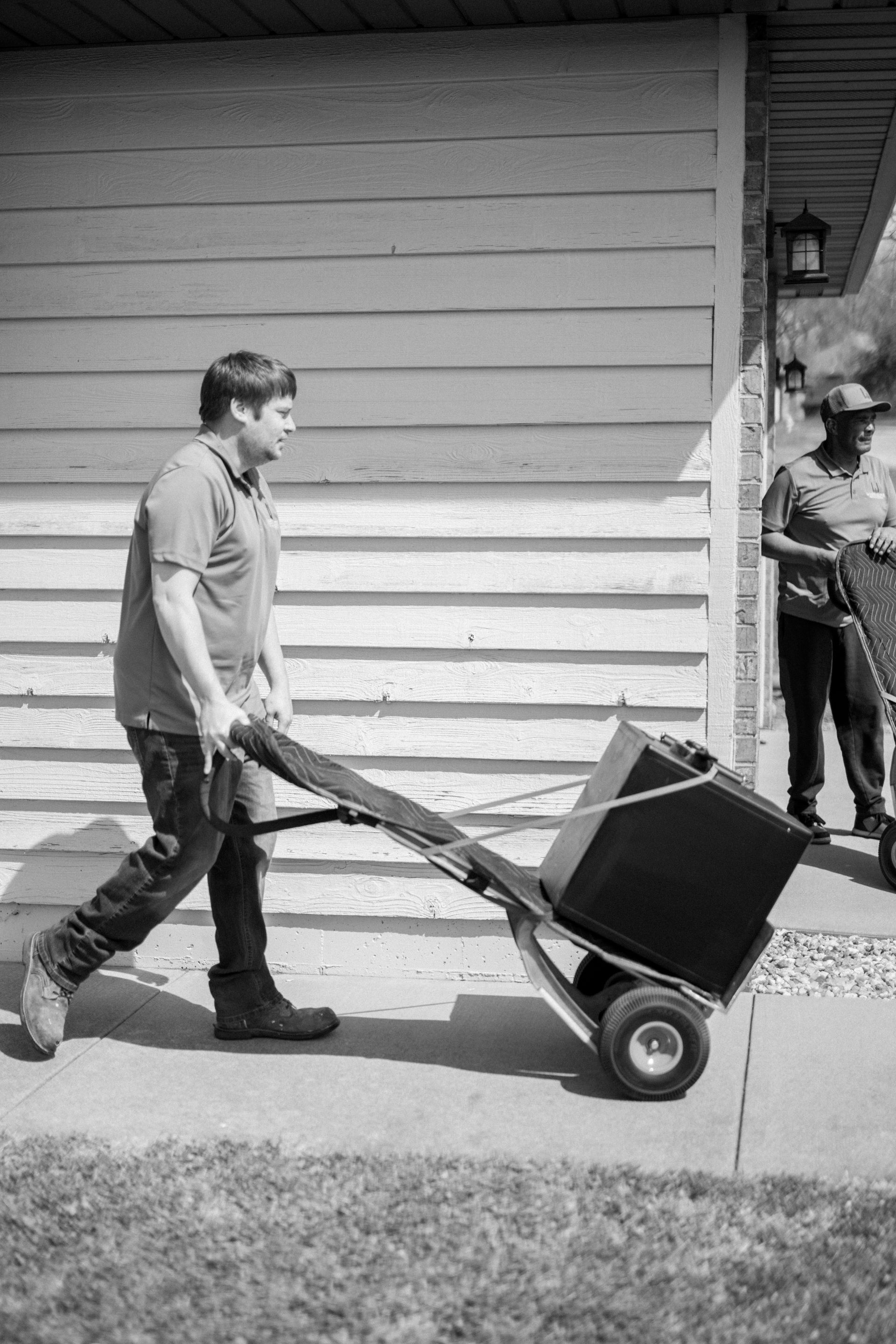 Man pushing a hand truck with a large box beside a white-sided building