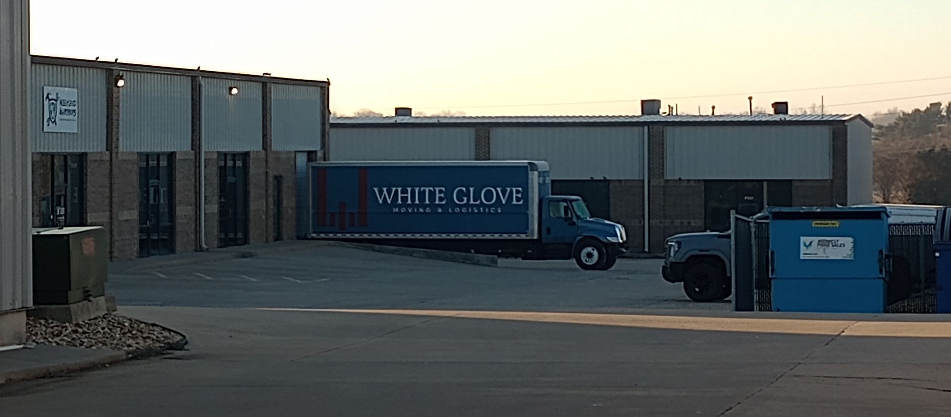 Industrial building entrance with a White Clove truck and a blue dumpster in the lot at dusk