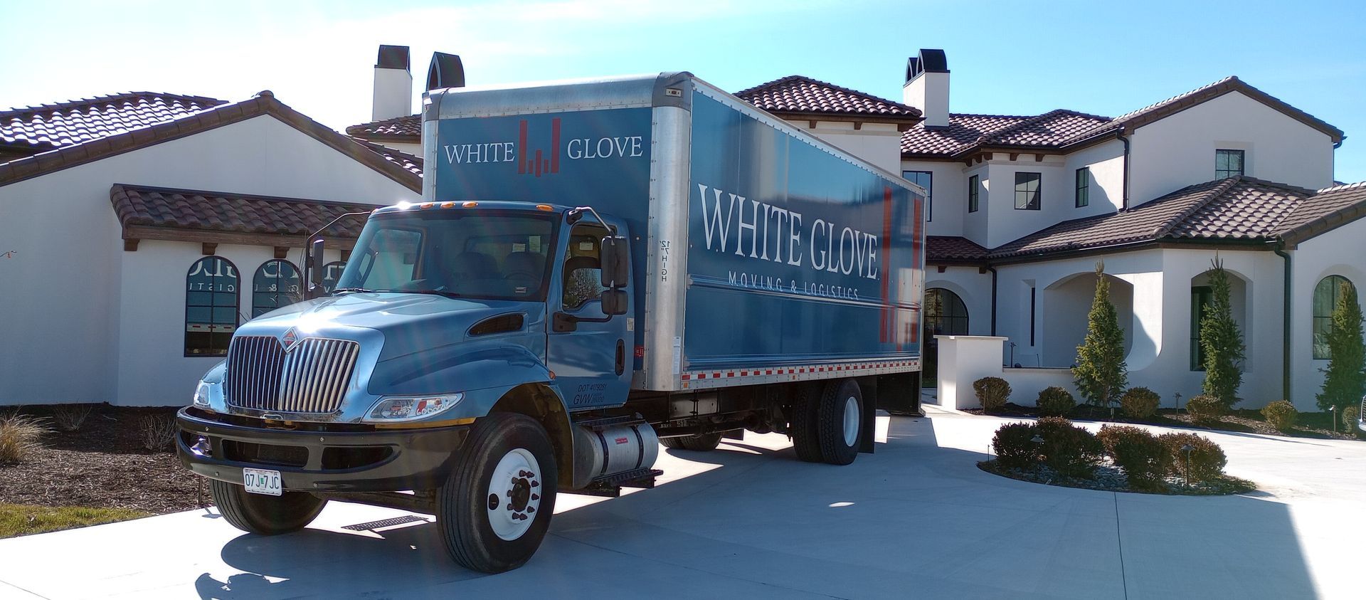 Blue moving truck parked in a driveway in front of a white house with a red-tile roof