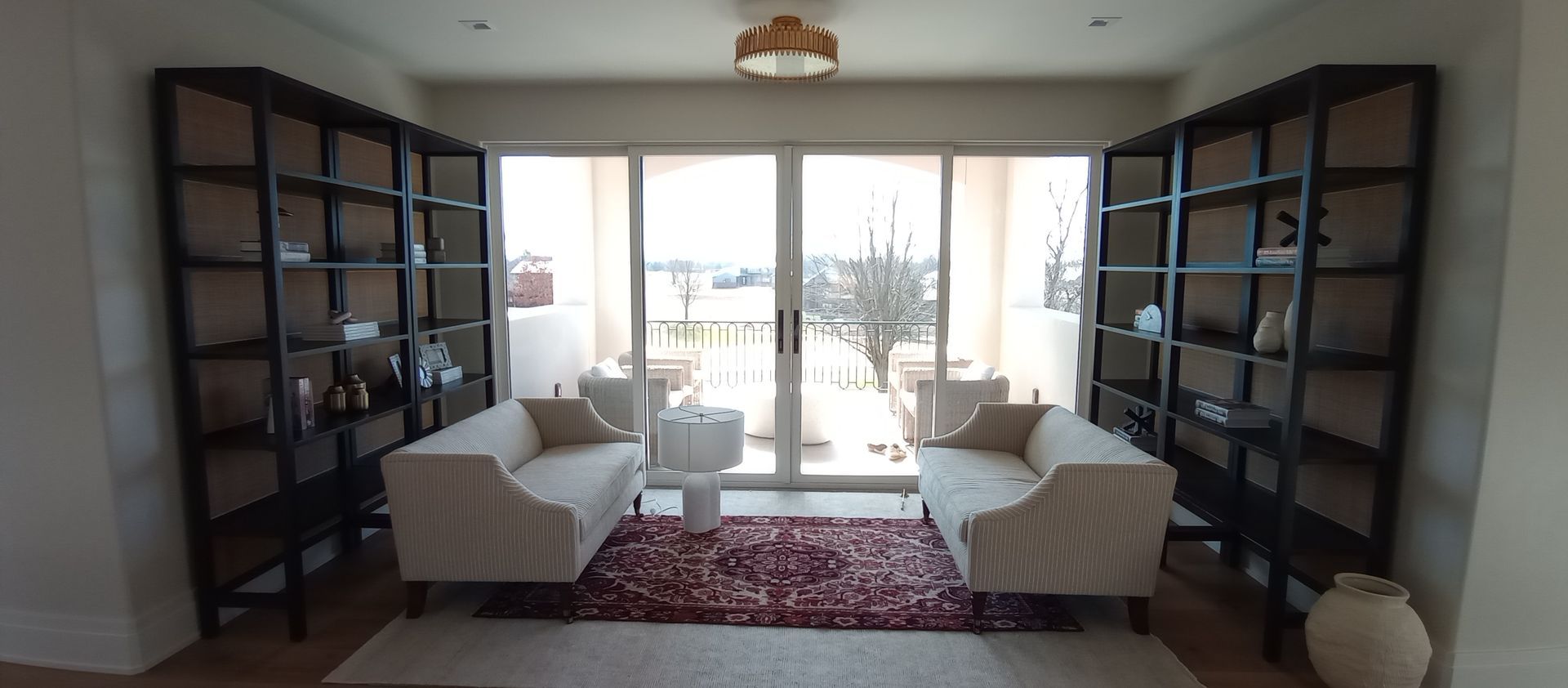 Bright living room with two white sofas, dark bookshelves, and glass doors opening to a balcony.
