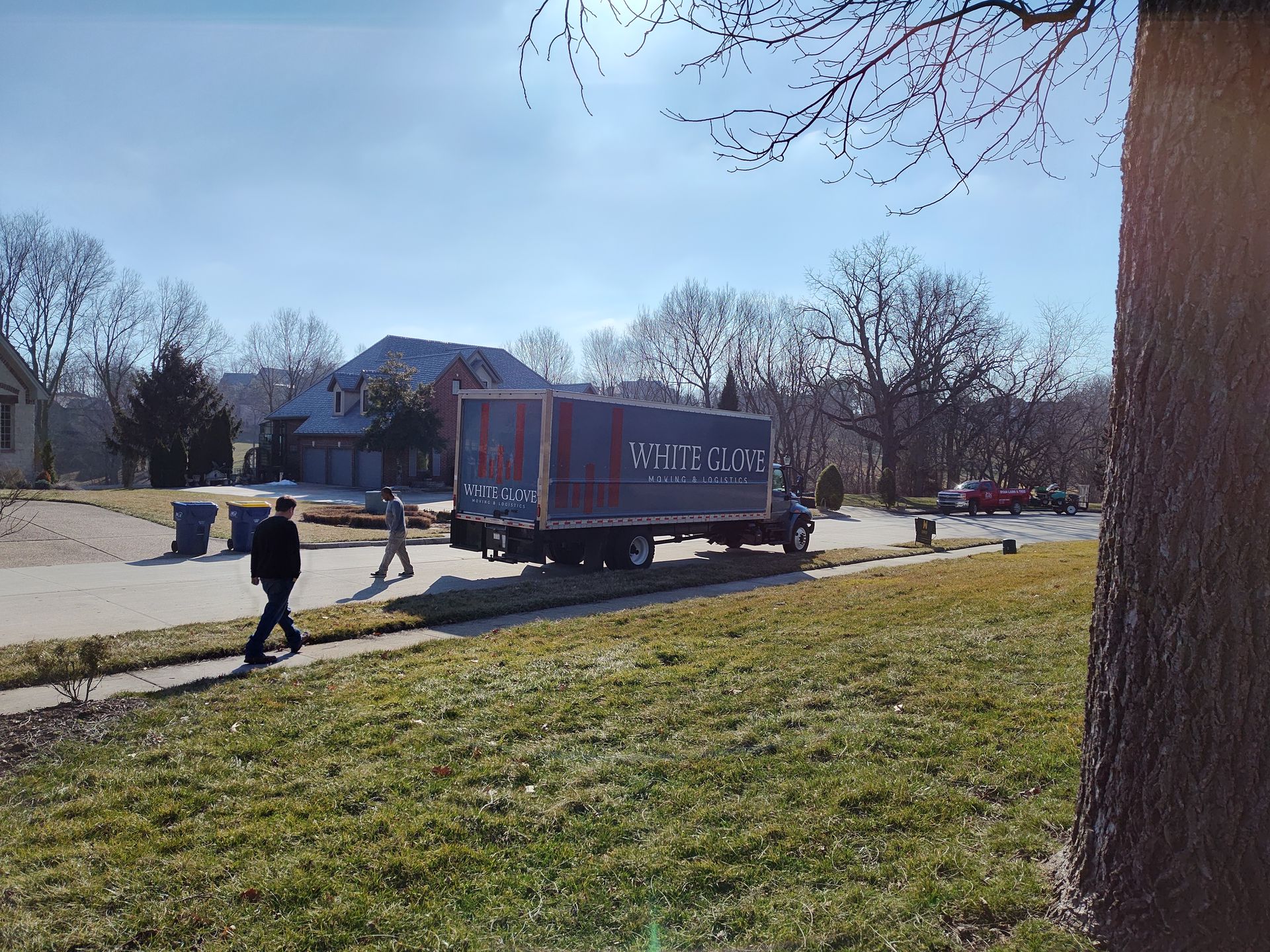 Delivery truck on a sunny suburban street with a person walking nearby and trash bins along the curb