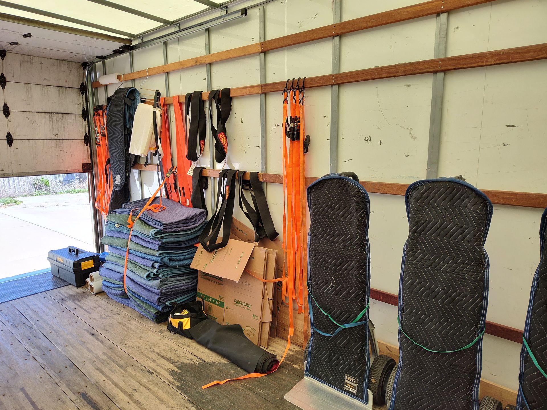 Truck bed with stacked mats, ropes, boxes, and orange tie-down straps inside a trailer or moving van.