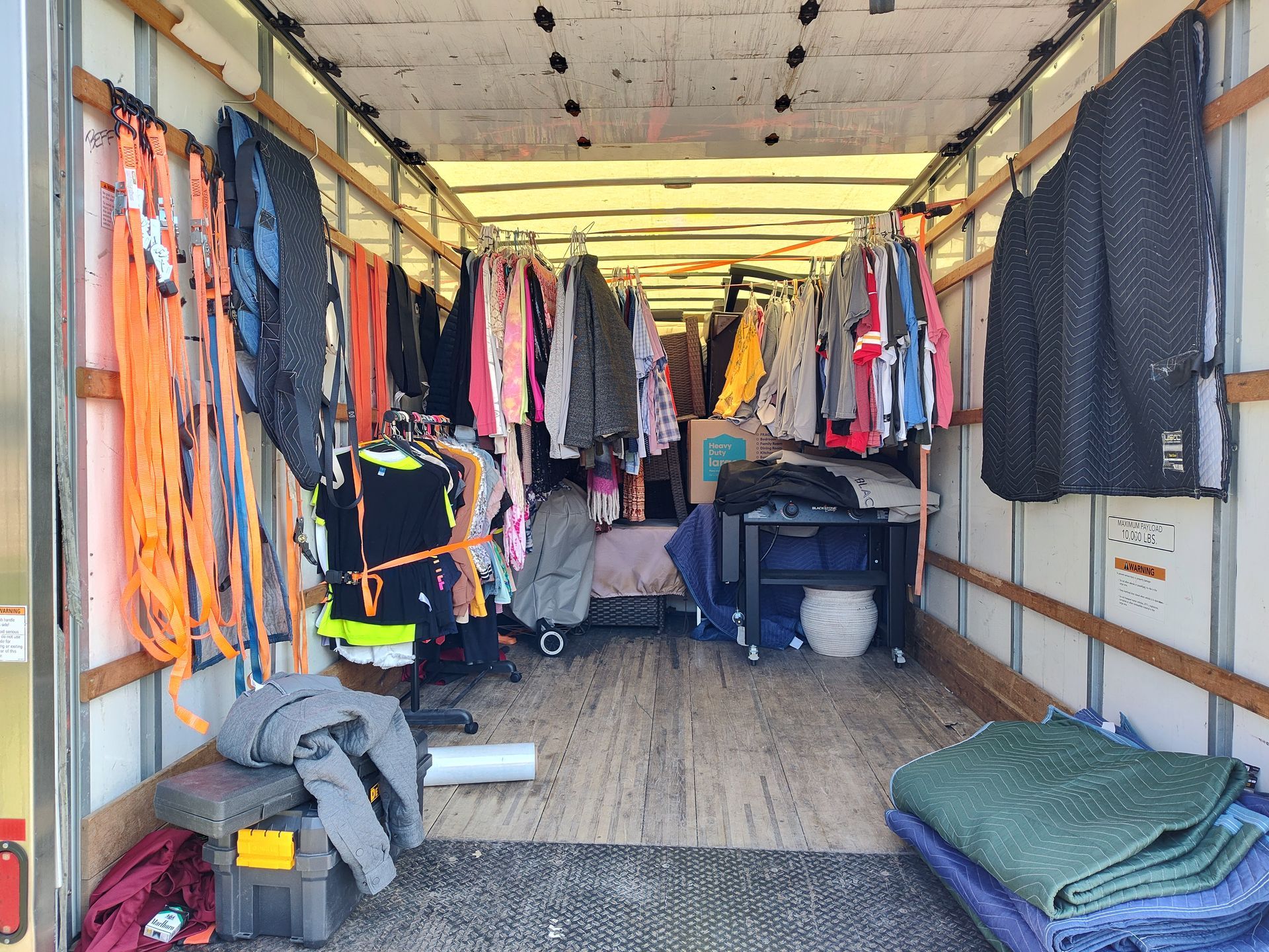 Inside a truck trailer, hanging clothes and storage bins line the walls with folded mats on the floor.