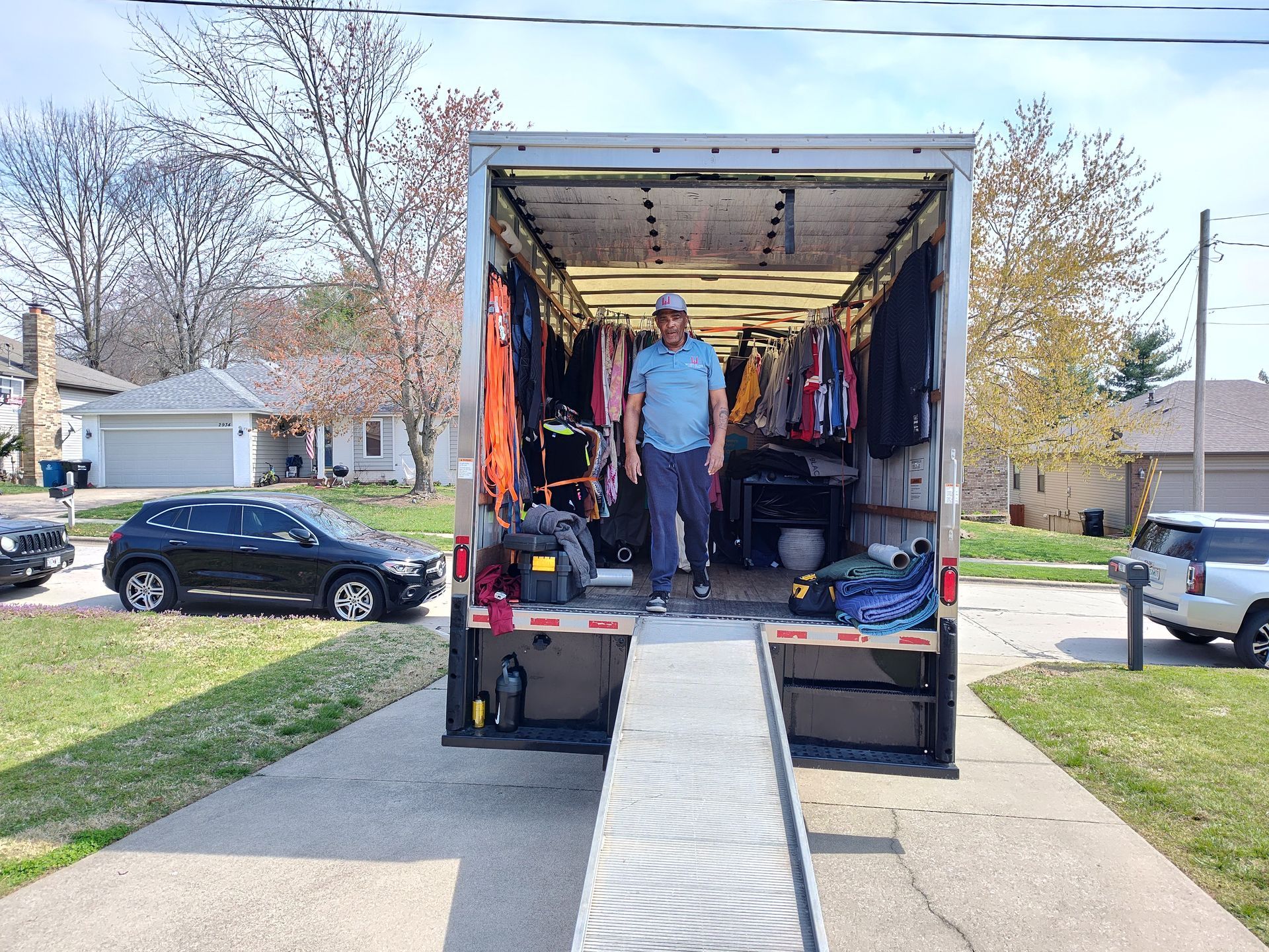 Person standing in a moving truck with a ramp extended to the sidewalk, parked on a residential street