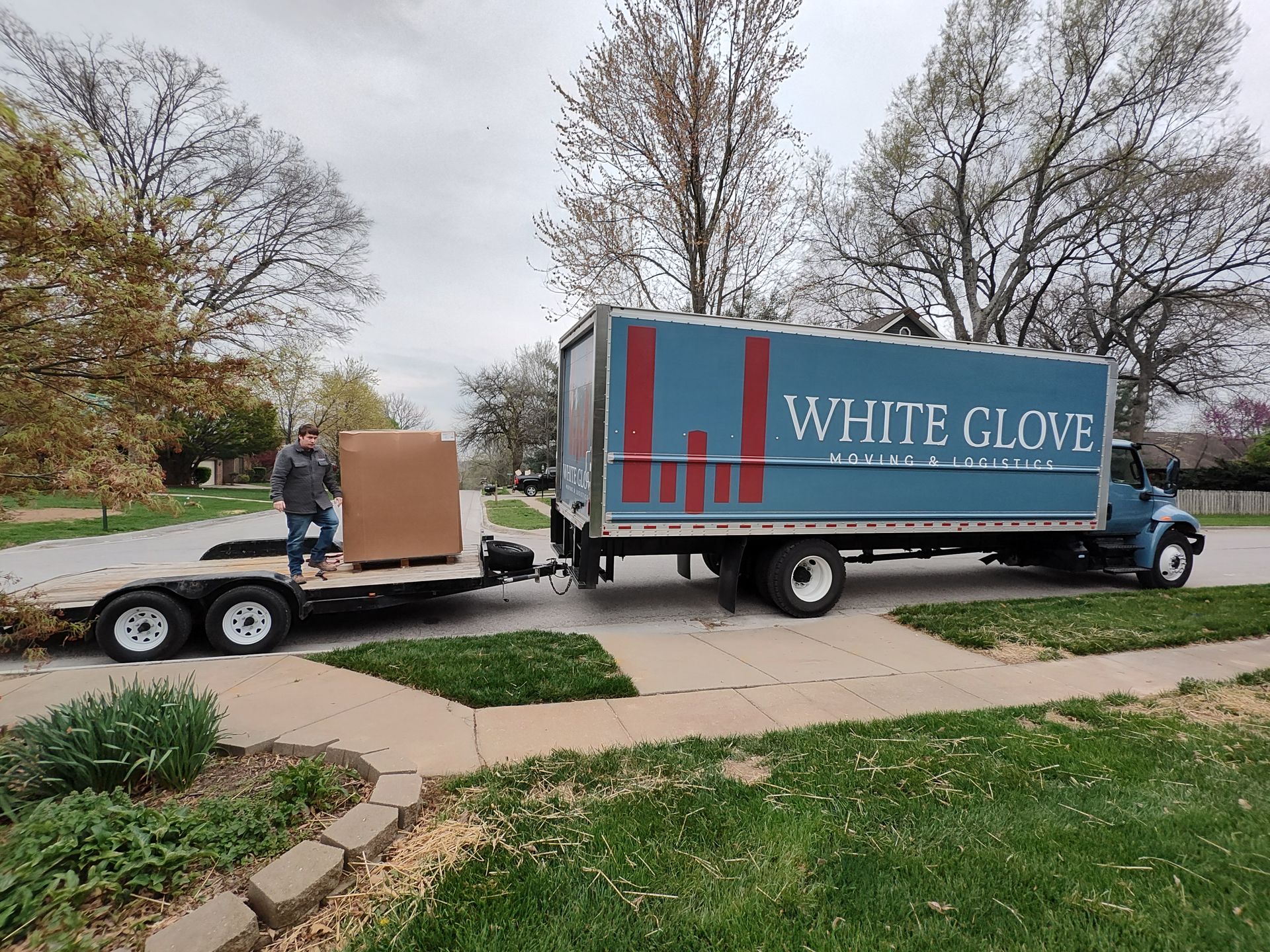 White Glove moving truck towing a trailer with furniture on a suburban street under cloudy skies