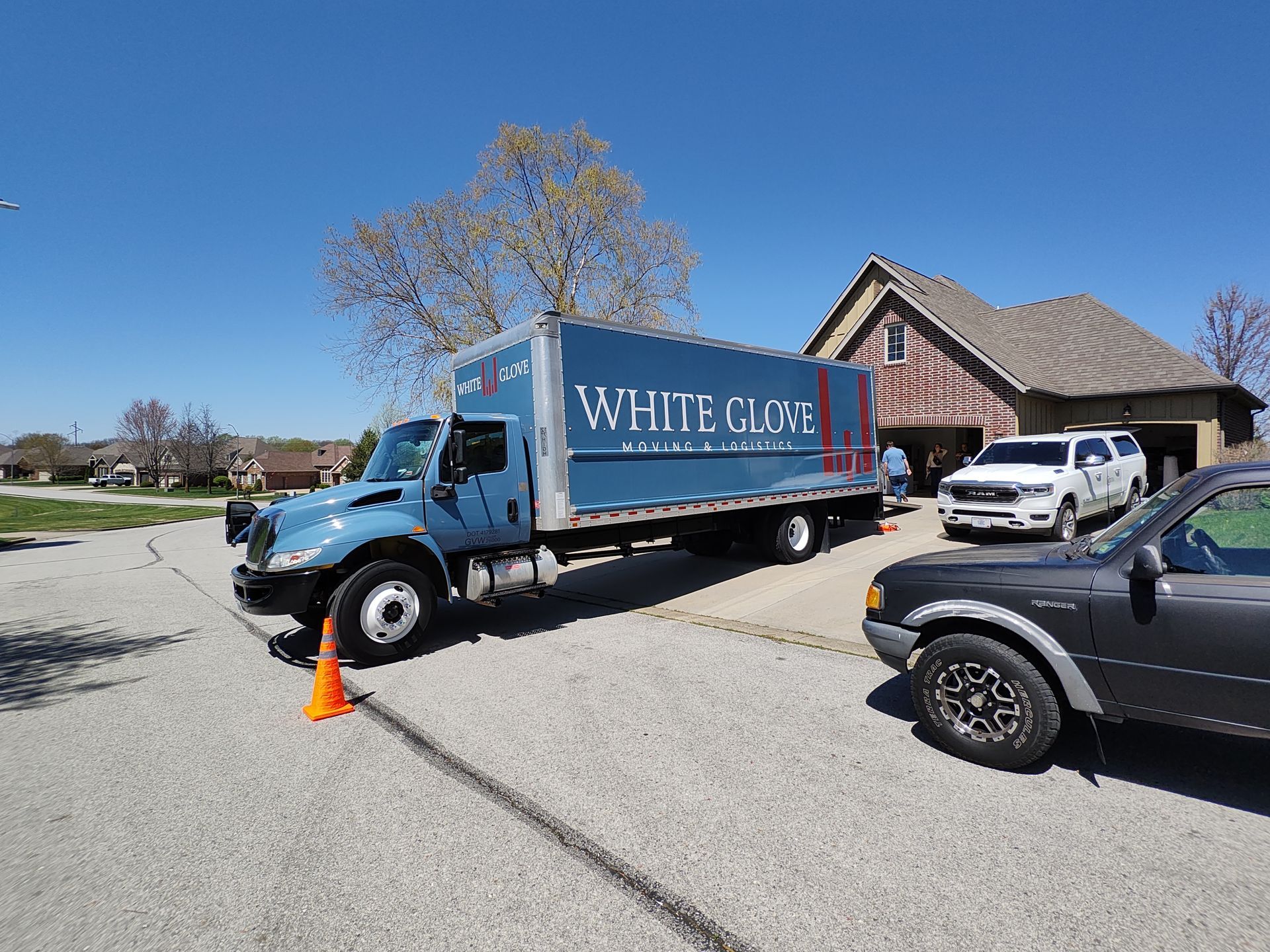 Blue moving truck parked by a building with a white SUV and black car nearby under a clear sky