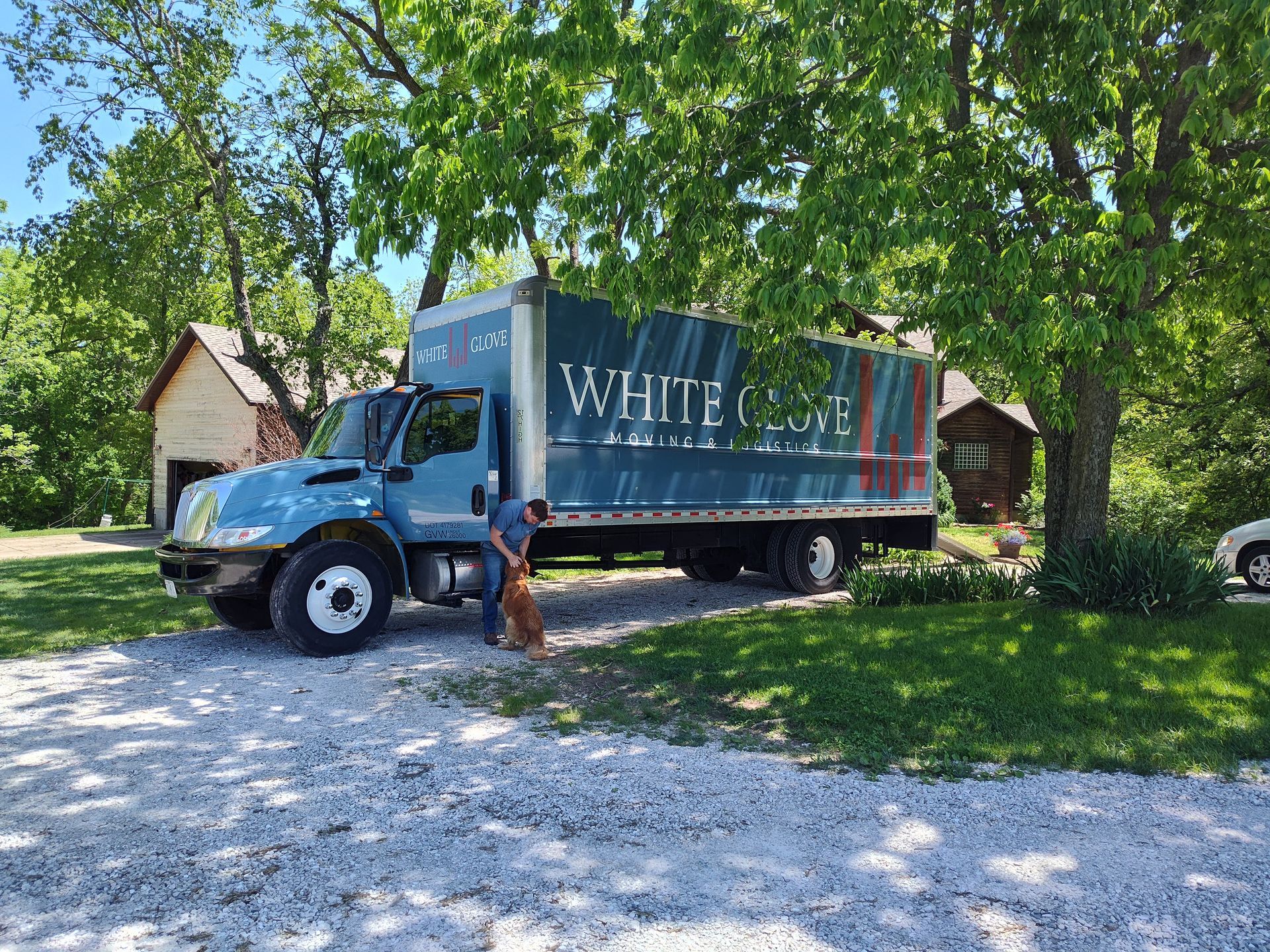 White truck with “White” logo parked under trees beside a house on a gravel driveway