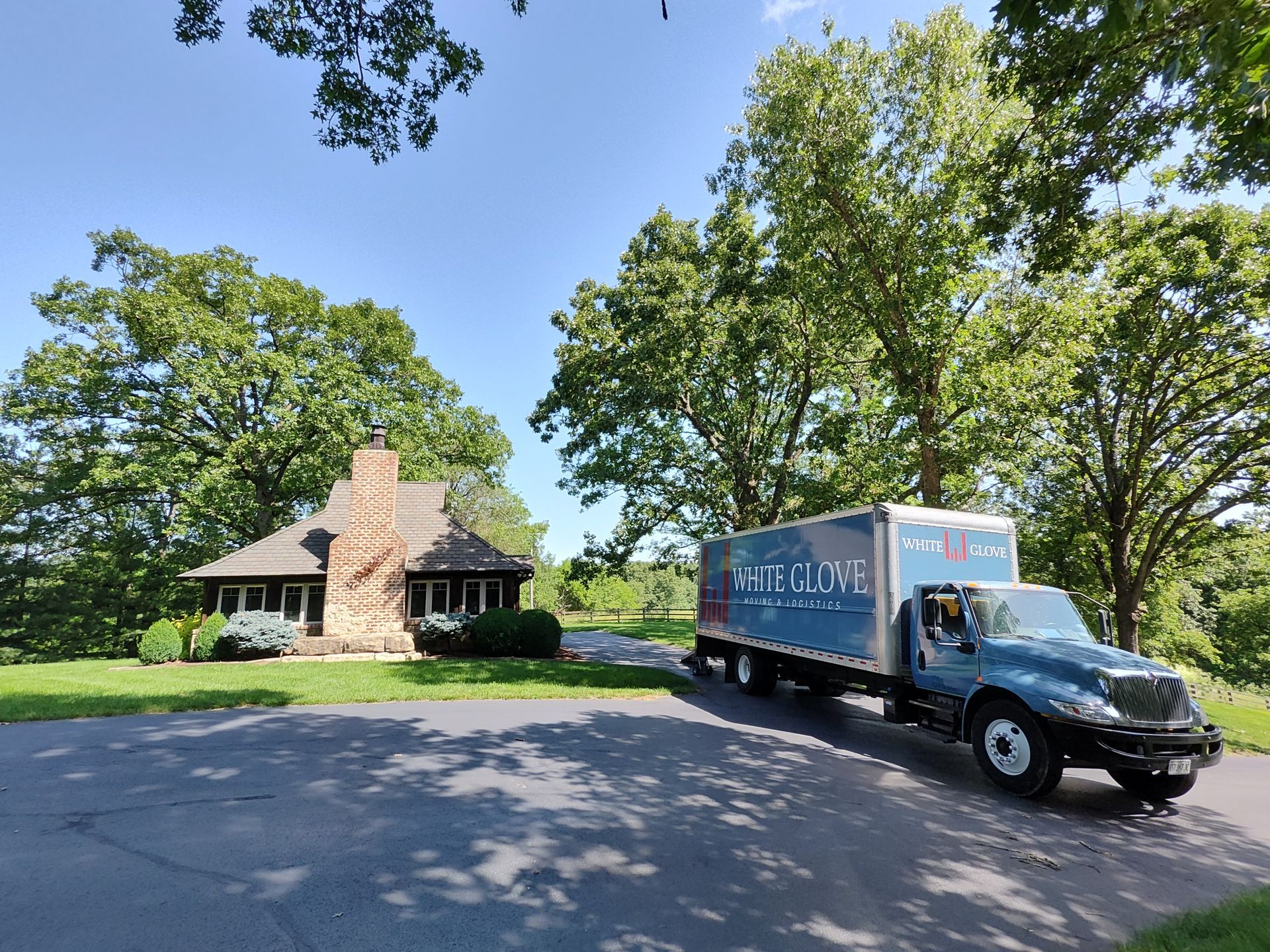 Moving truck parked on a driveway beside a stone house and green trees on a sunny day