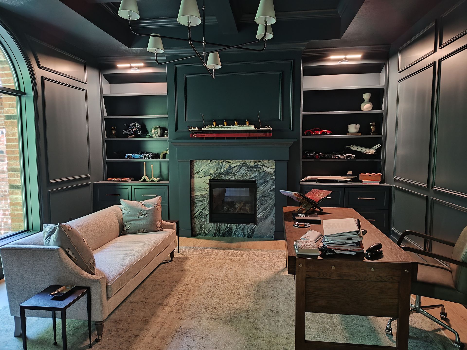 Cozy dark-paneled living room with a fireplace, sofa, desk, bookshelves, and large window