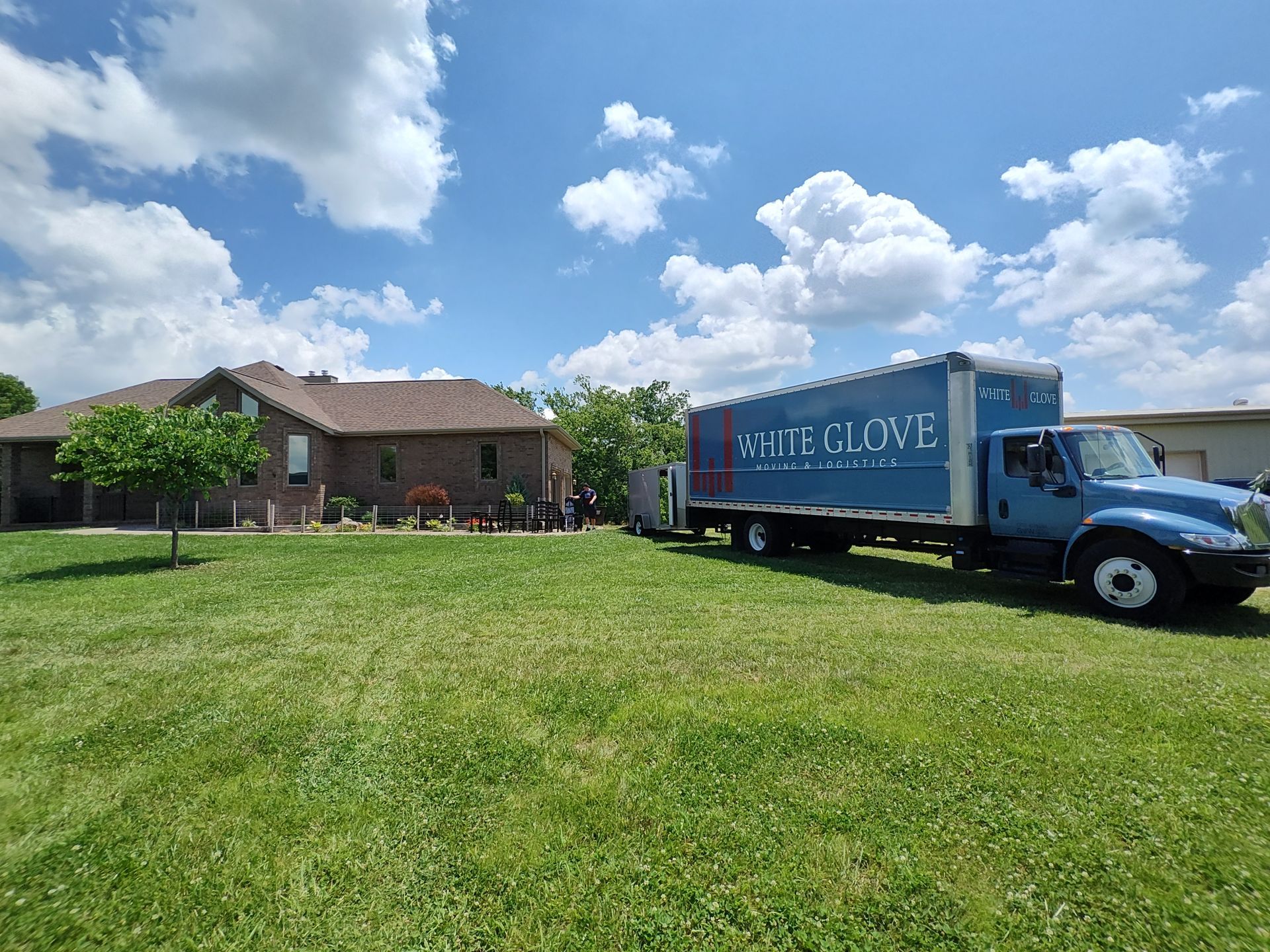 Blue moving truck parked beside a brick house on a sunny lawn under a partly cloudy sky