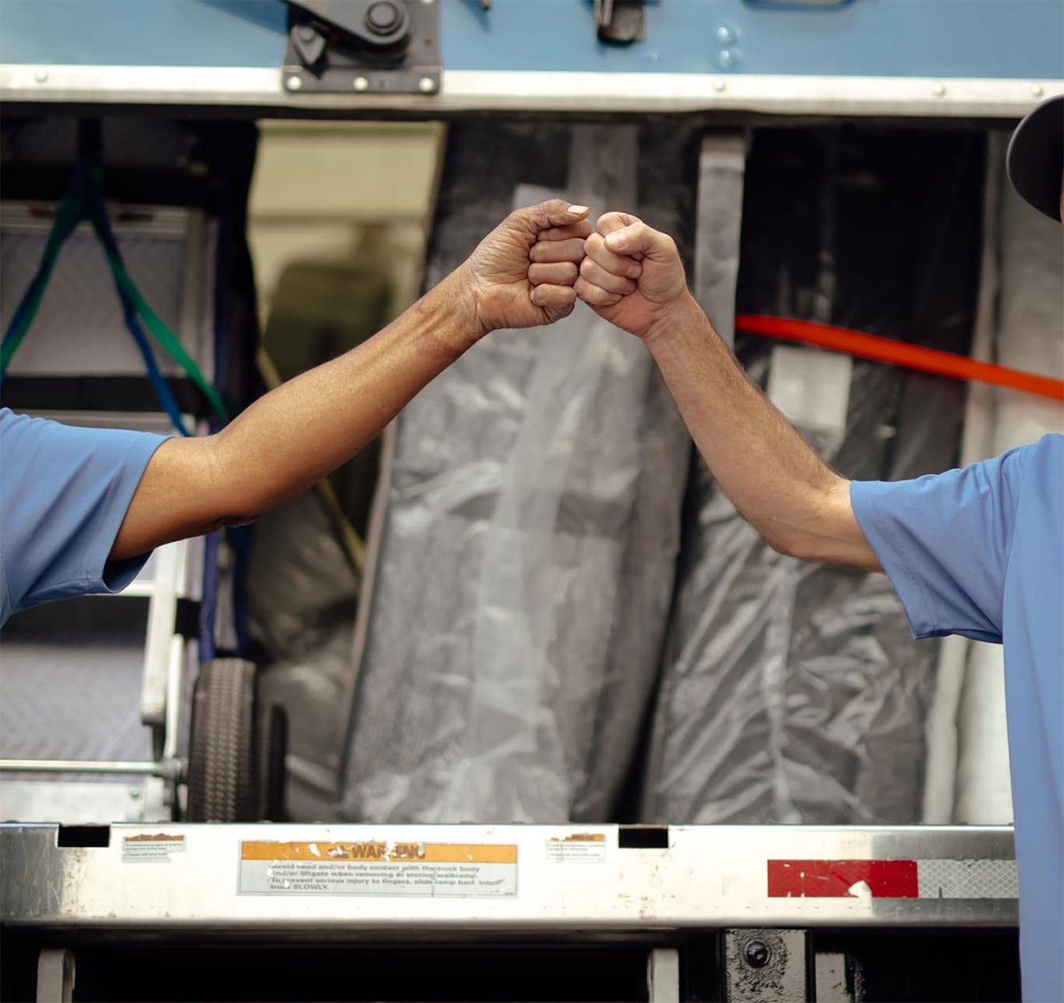 Two workers in blue shirts fist bump across a conveyor belt in an industrial setting