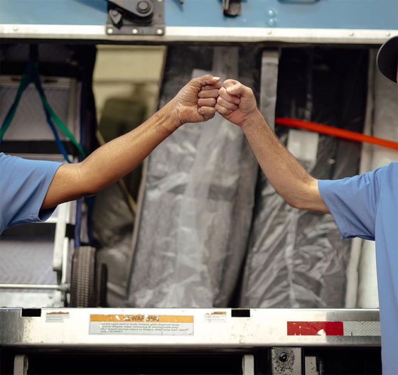 Two workers in blue shirts fist bump across a conveyor belt in an industrial setting