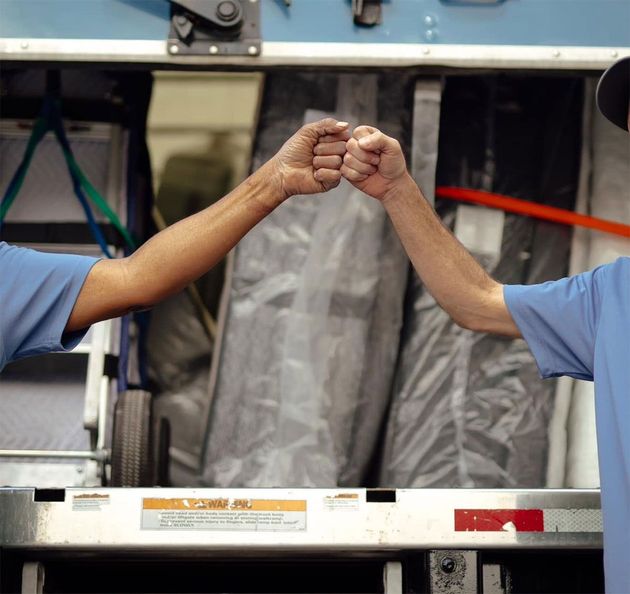 Two workers in blue shirts fist bump across a conveyor belt in an industrial setting