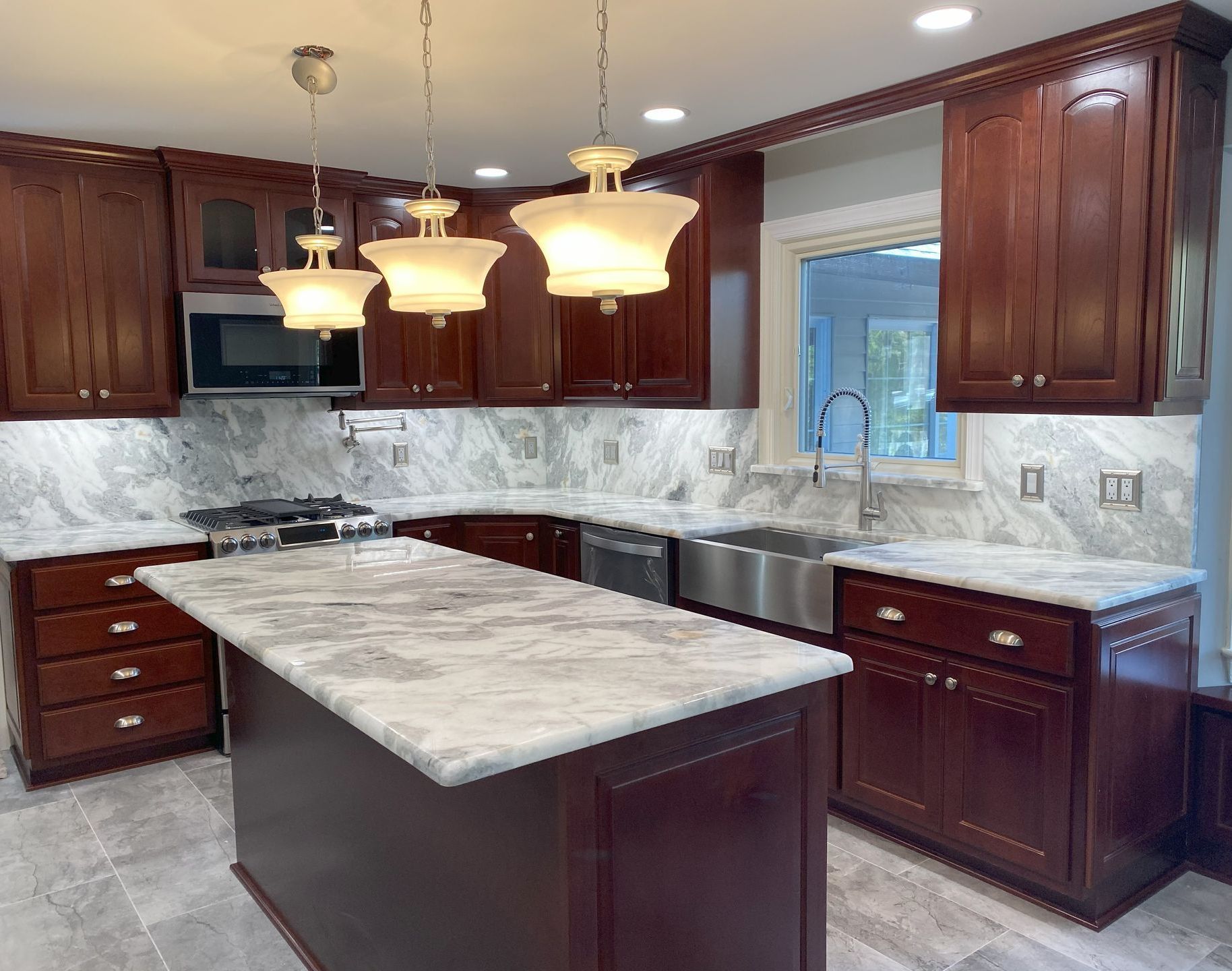 A kitchen with dark wood cabinets, marble countertops, stainless steel appliances, and island with pendant lights.