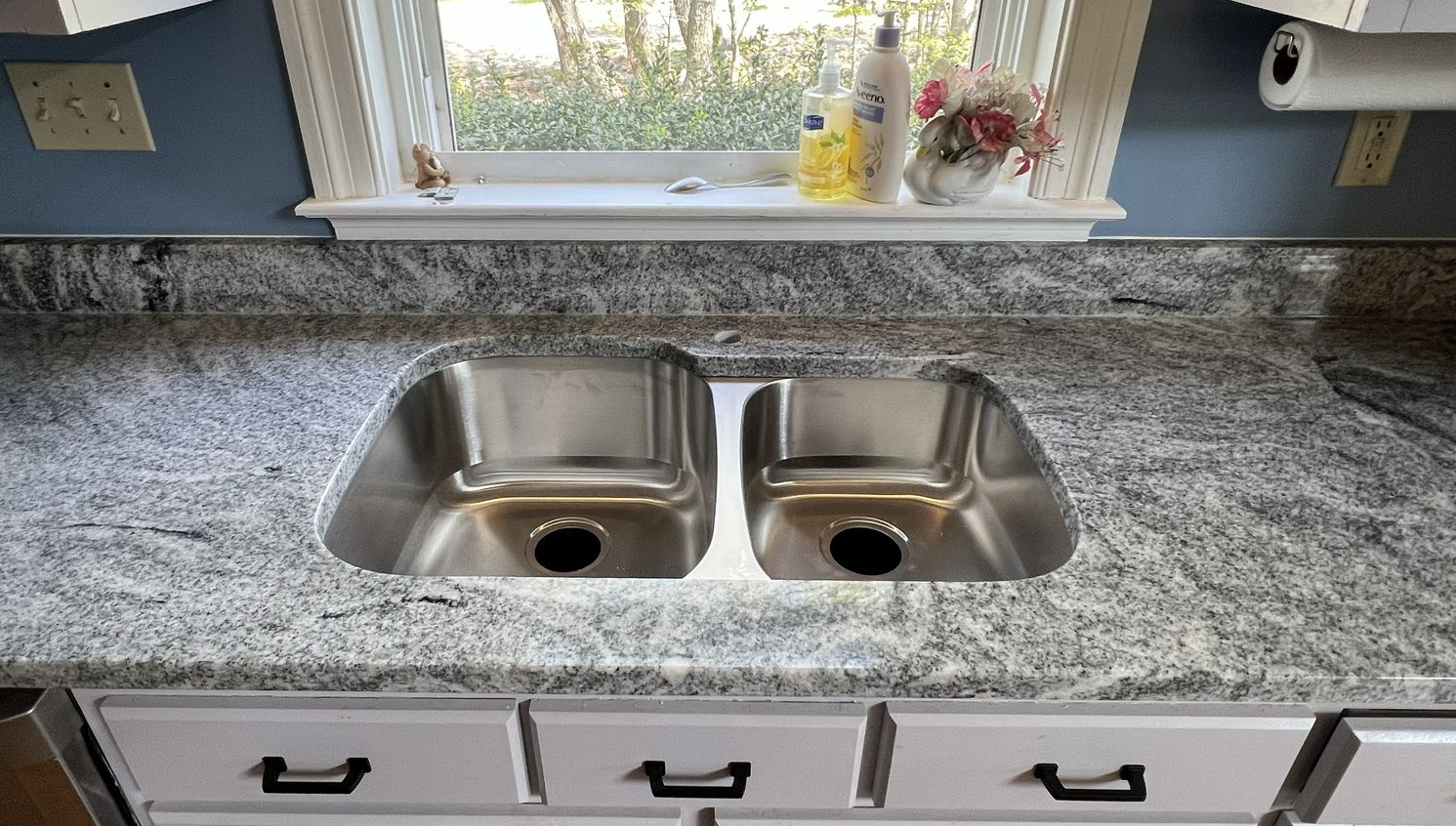 Kitchen countertop with double stainless steel sink, window above. Granite-like surface, white cabinets.
