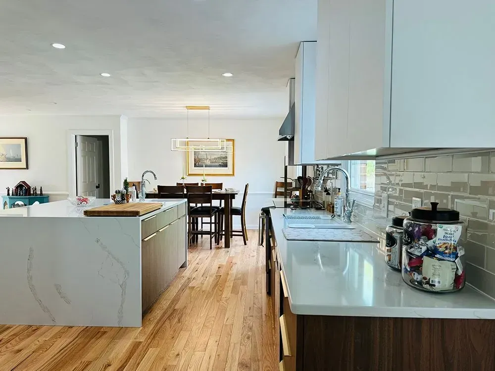Modern kitchen with white cabinets, wood floors, and island. Dining area in the background.