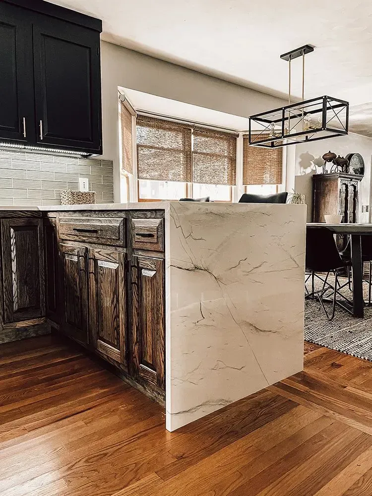Kitchen island with rustic wood cabinets and light-colored countertop.  Black cabinets and a window with blinds.