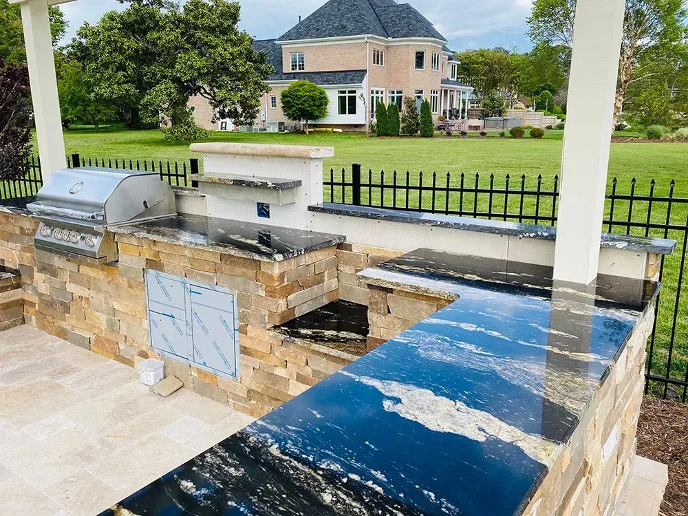 Outdoor kitchen with stone counters, grill, and view of a large house and green lawn.