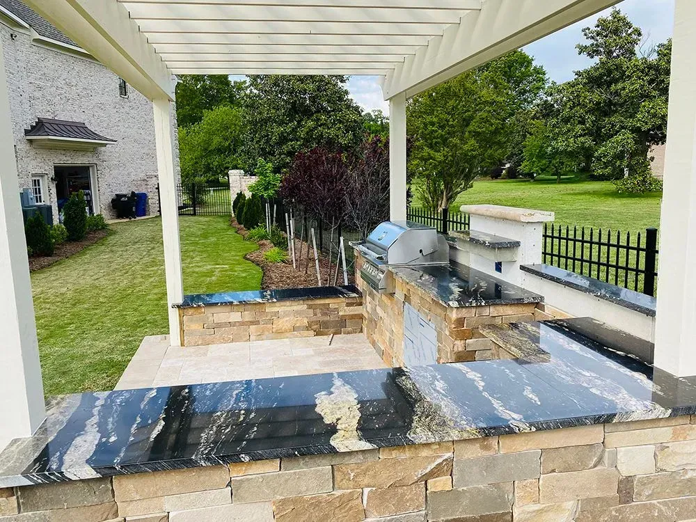 Outdoor kitchen with granite countertop and grill, under a pergola, adjacent to a green yard and house.