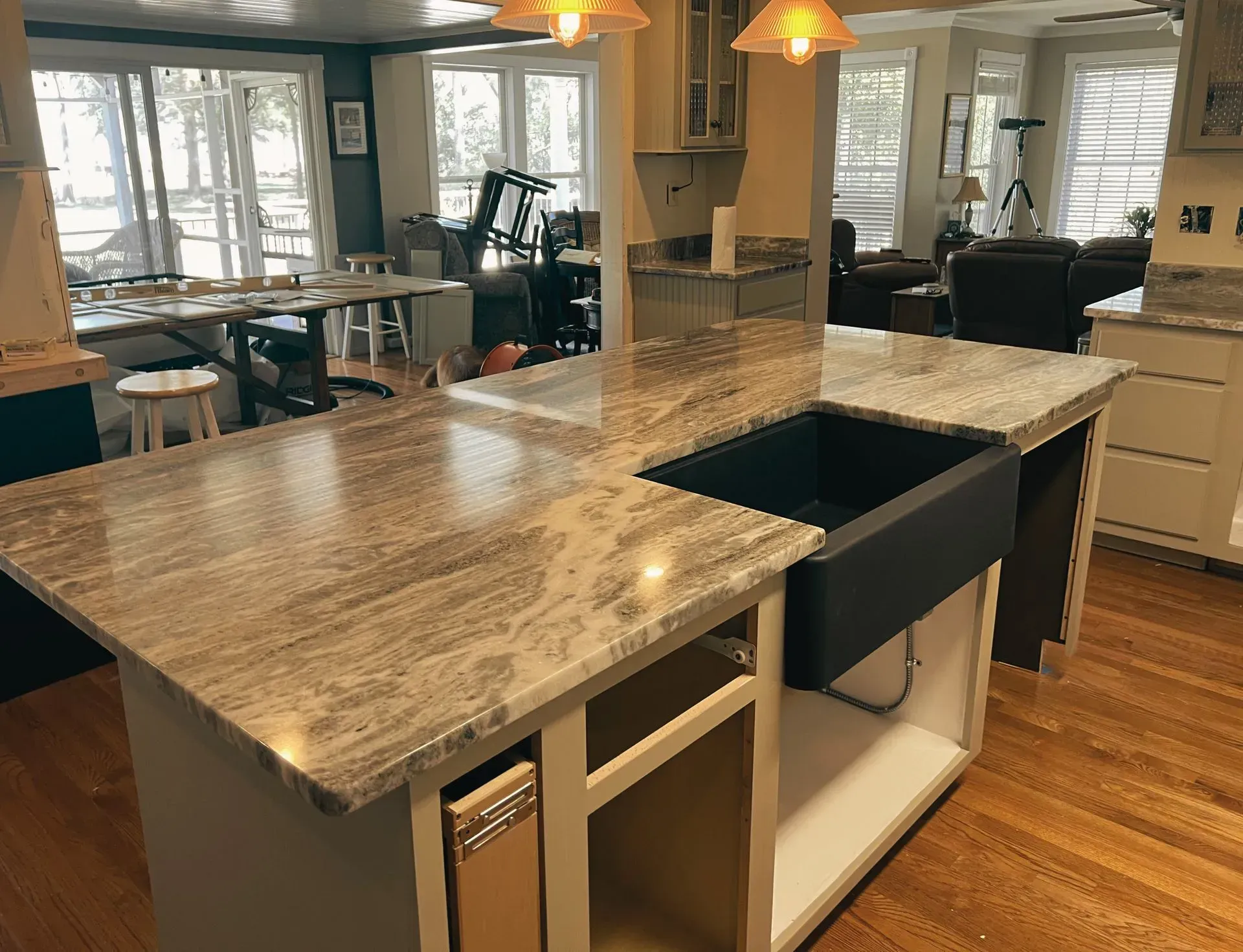 Kitchen island with granite countertop, black farmhouse sink, and open cabinetry. Wooden floor, large windows.