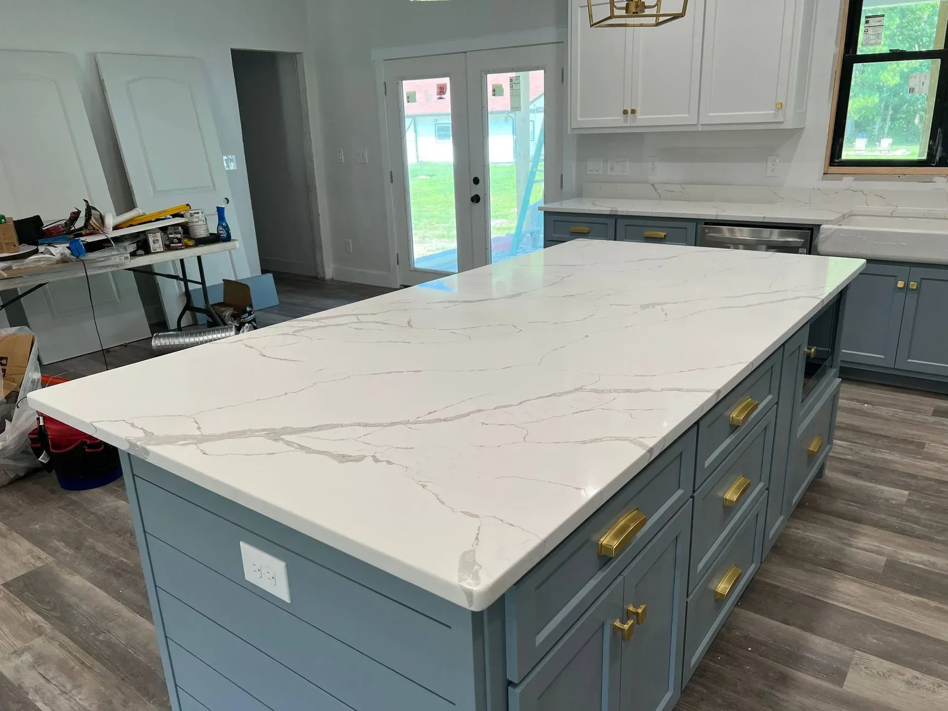 Blue kitchen island with white countertop, brass hardware. Cabinets and French doors in background.